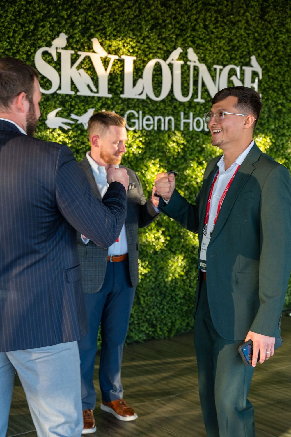 Business professionals in suits networking and conversing at an upscale event with green hedge wall backdrop.