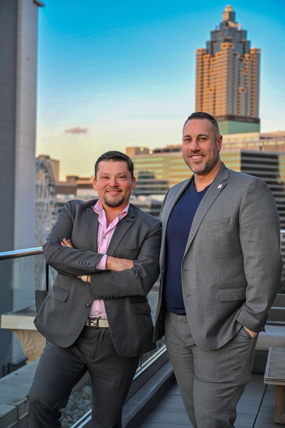Two professionals in business attire posing on a rooftop terrace with city skyline at sunset.