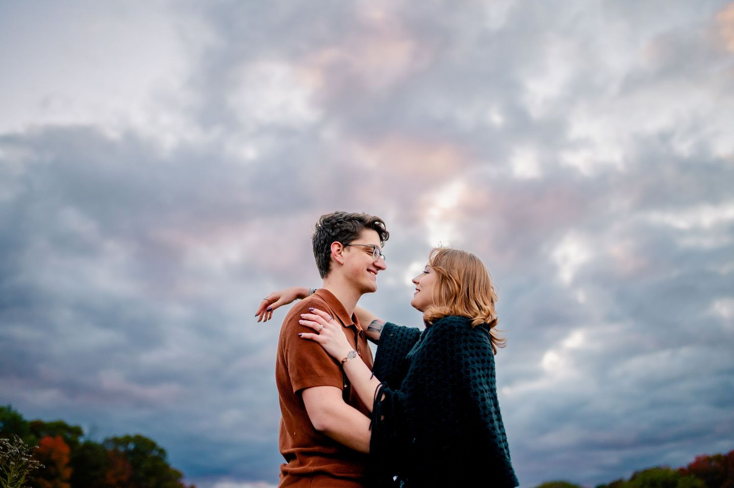 A romantic couple embraces under dramatic cloudy skies at sunset in a natural outdoor setting.