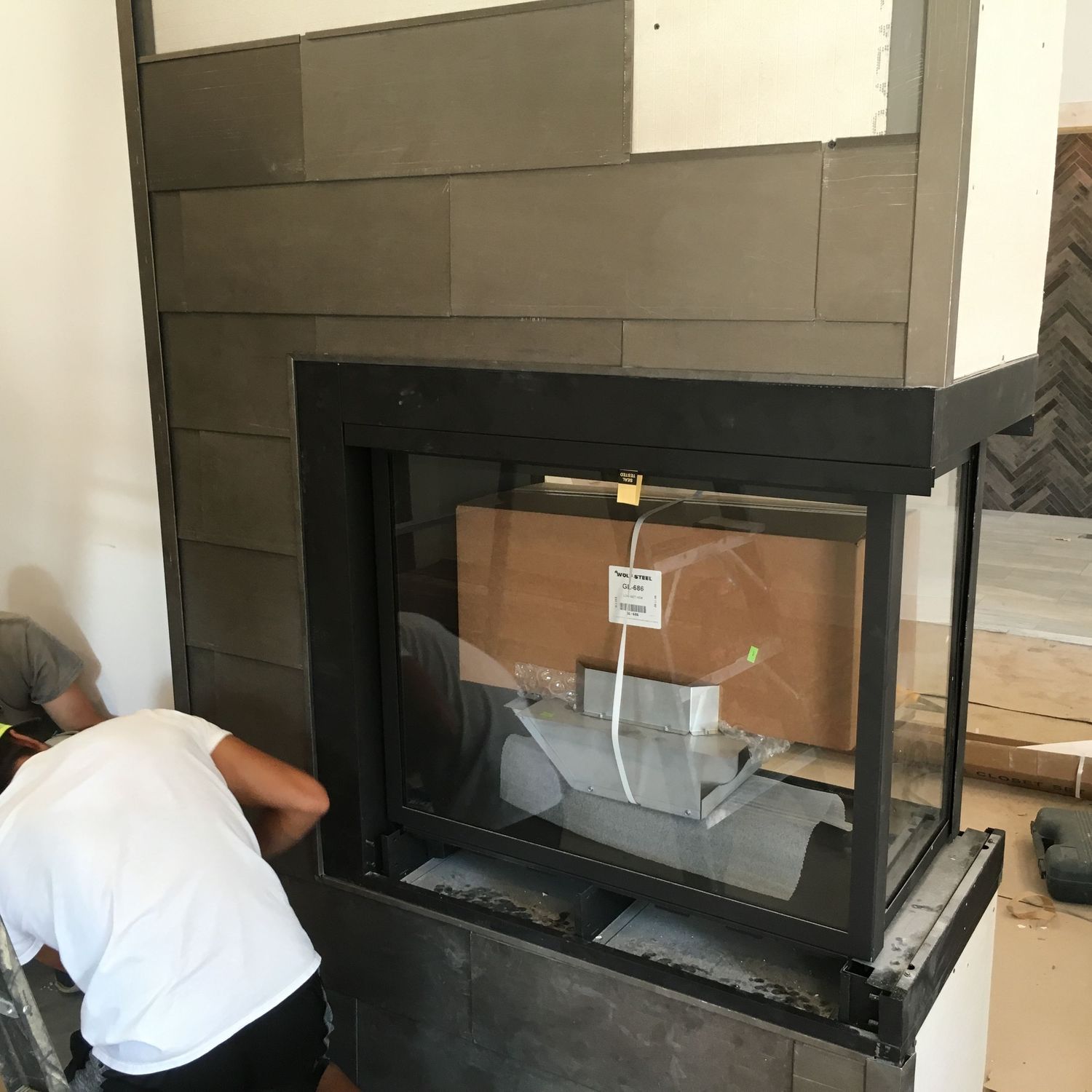 A modern three-sided fireplace installation in progress with black tile surround and cardboard protective covering.