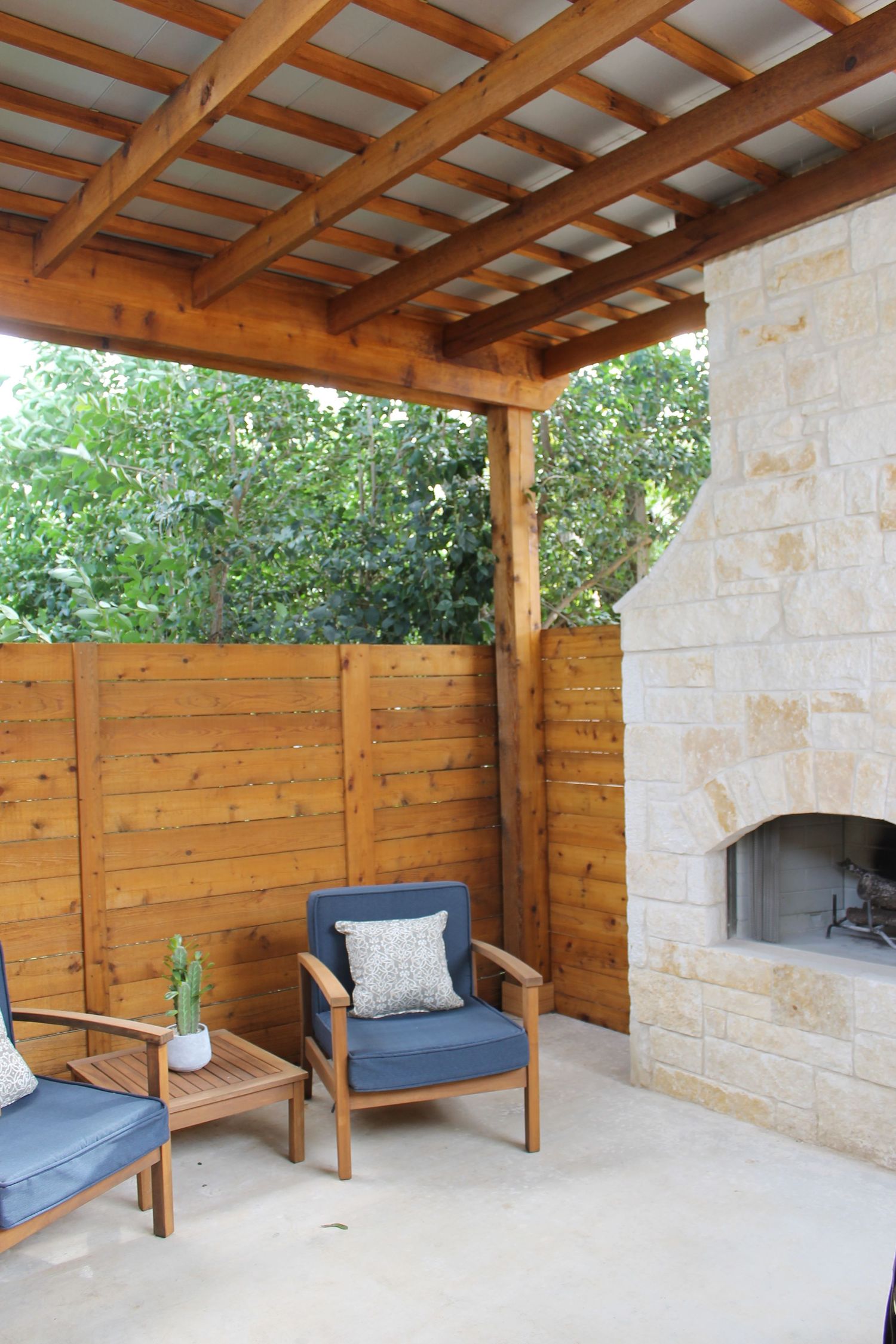 Modern outdoor patio with wooden pergola, stone fireplace, and teak lounge chairs featuring blue cushions and privacy fence.