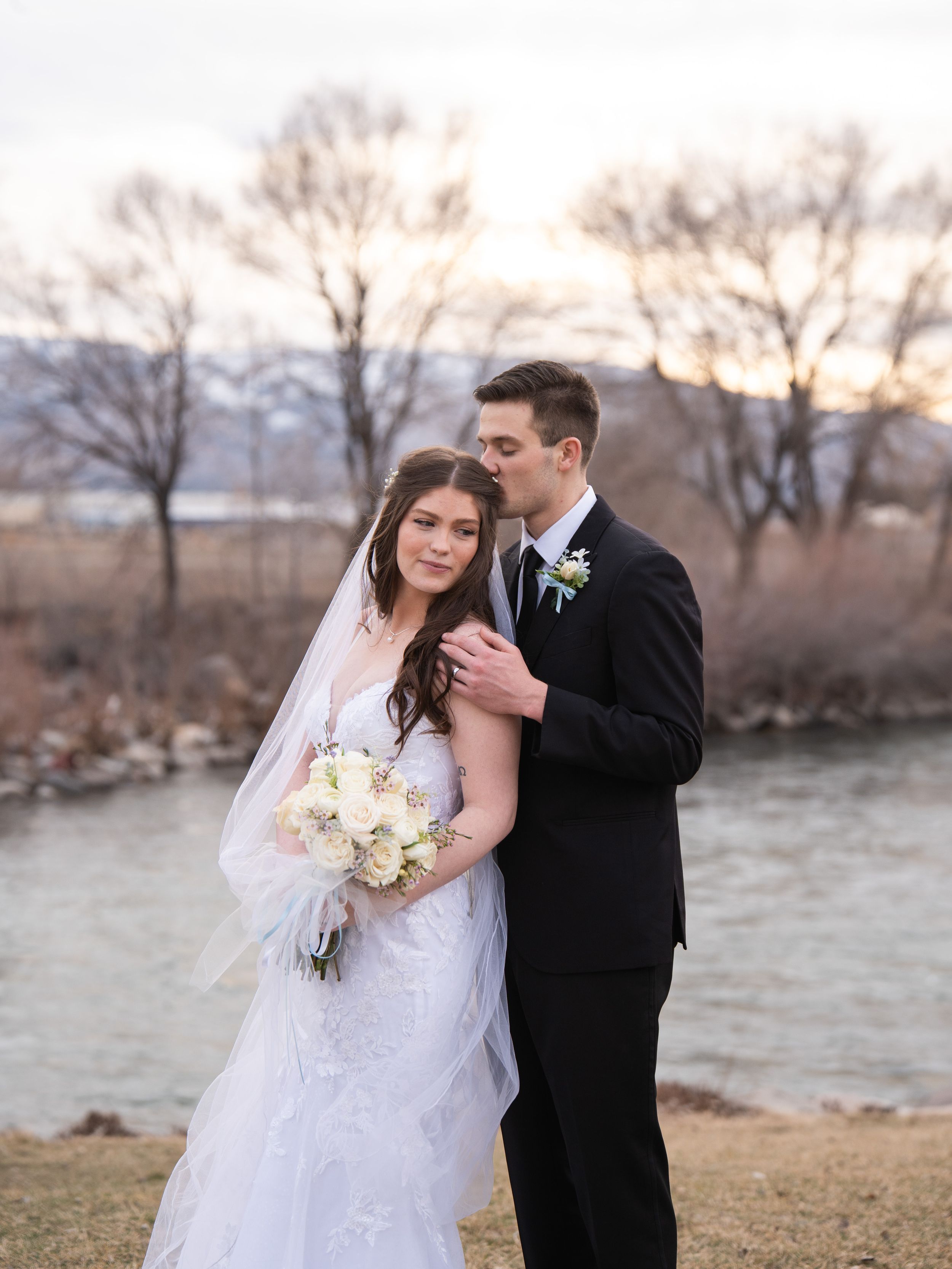 A romantic winter wedding couple embraces by a river at sunset in elegant formal attire.