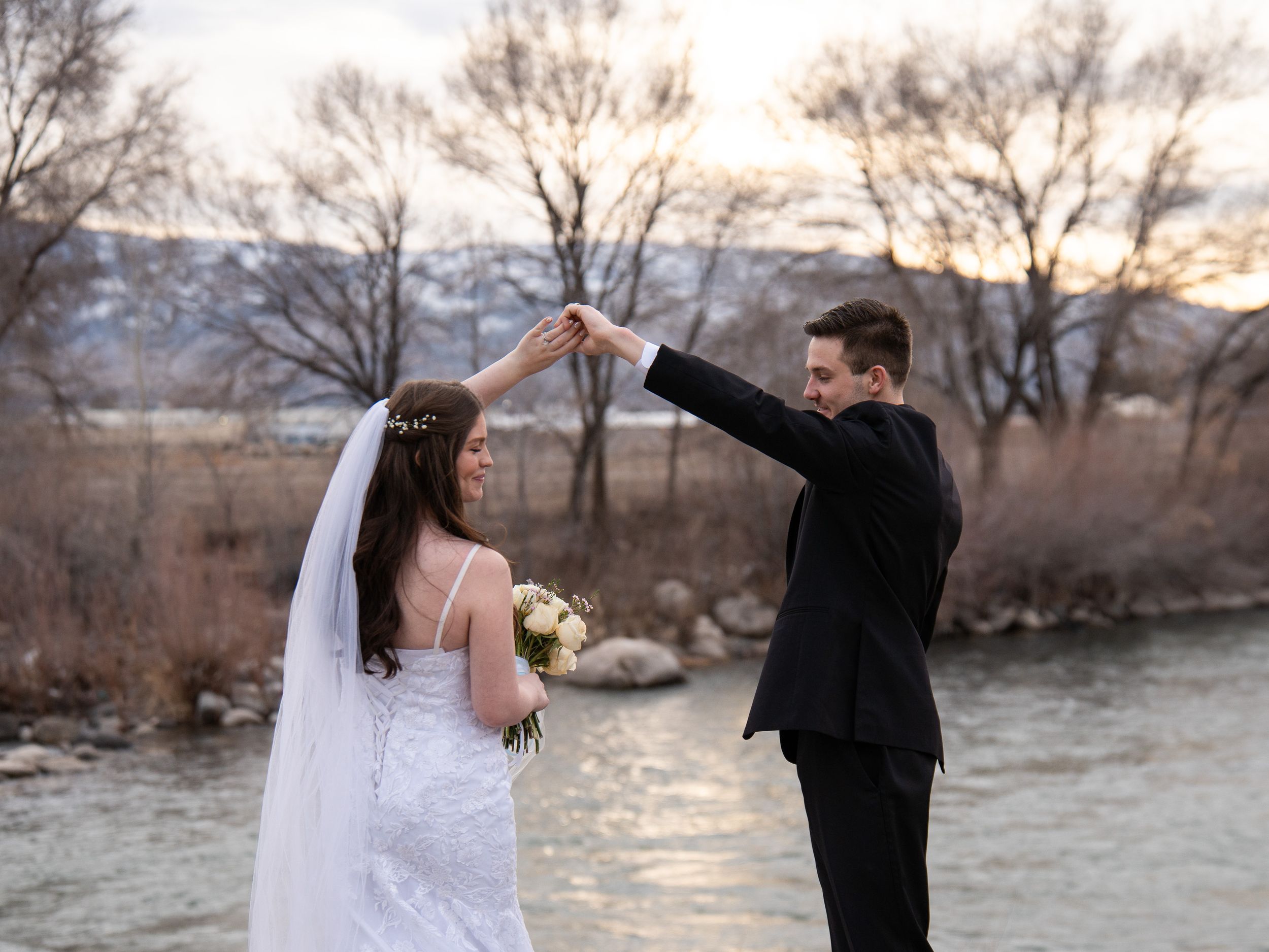 A couple in wedding attire dances together at sunset by a river with mountains in the background.
