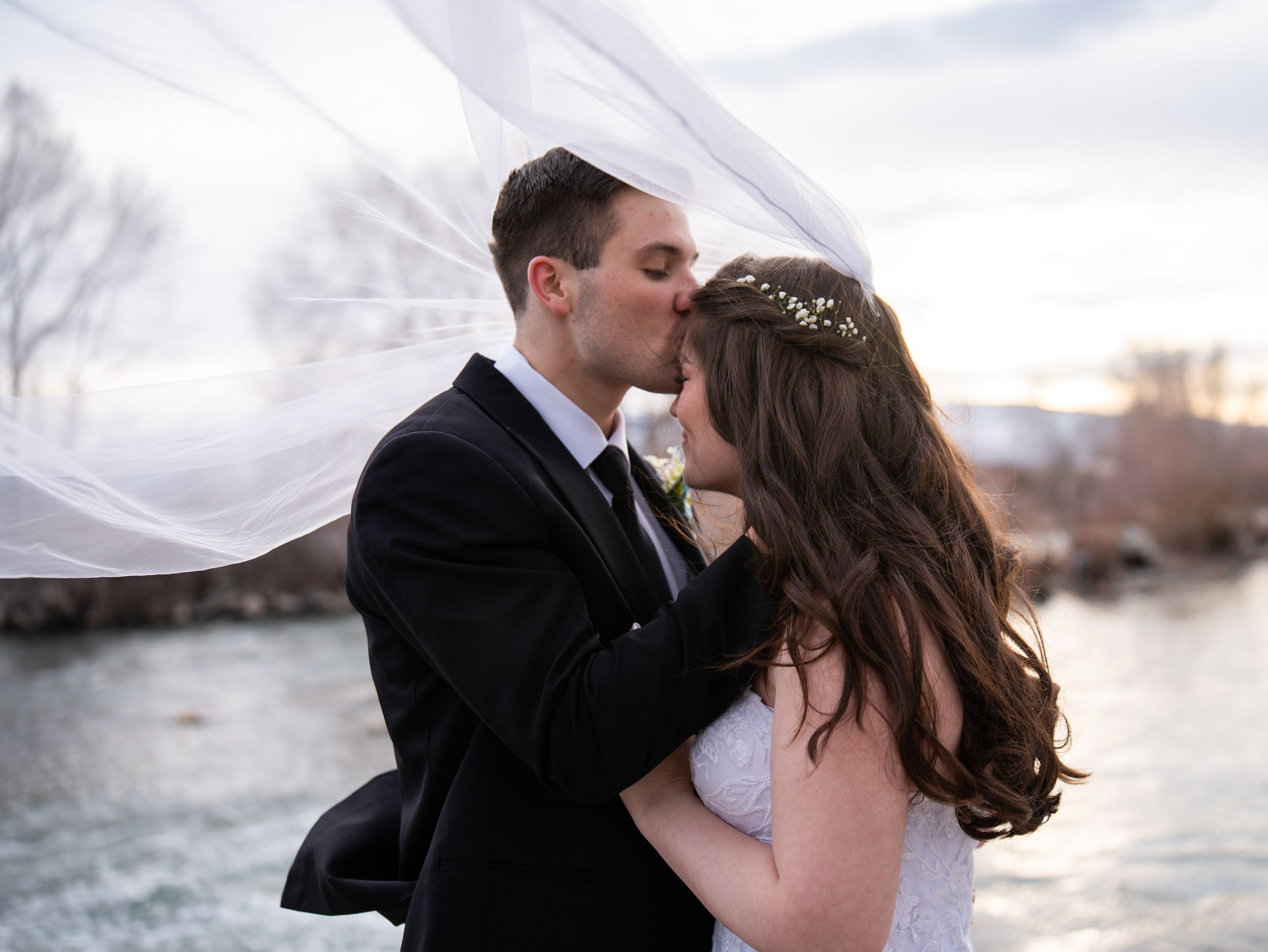 A romantic couple in formal wedding attire embraces and kisses by a serene waterfront at sunset.