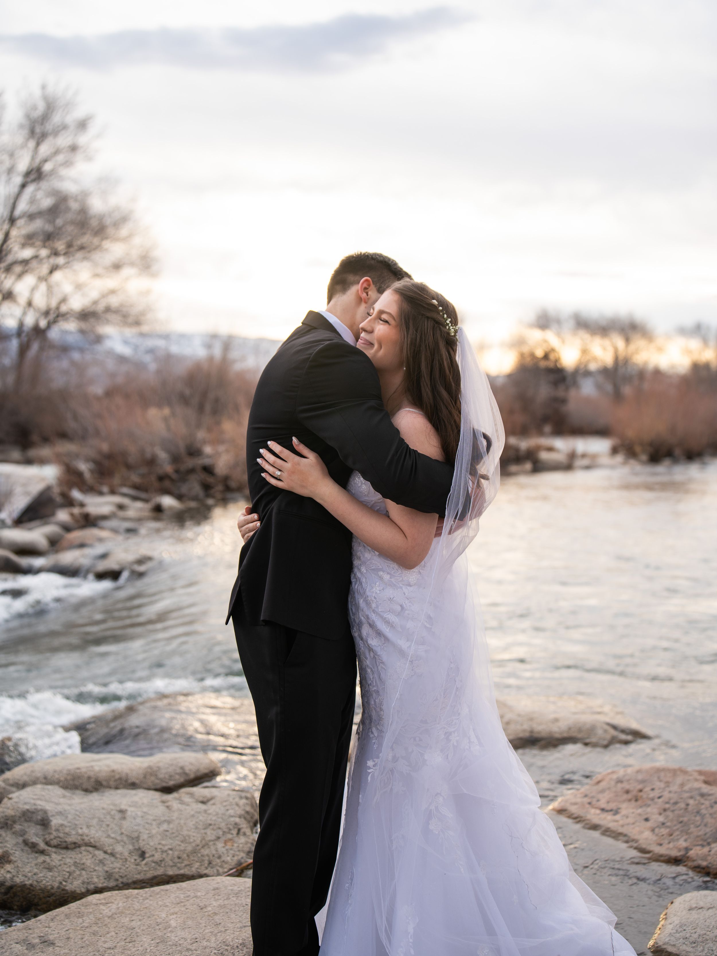 Romantic couple embraces by a river during sunset with snowy mountains and bare trees in the background.