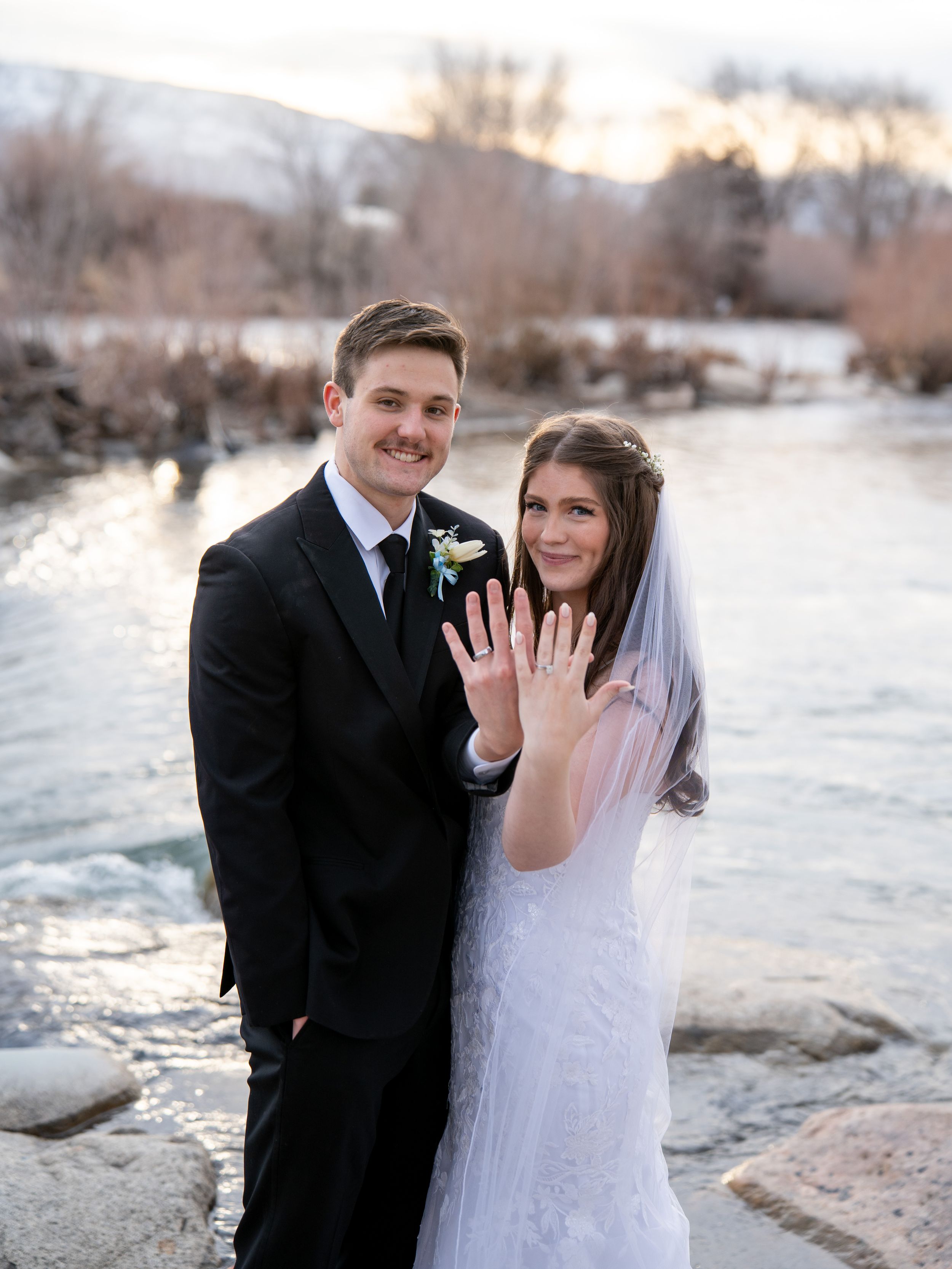 A couple in winter wedding attire poses on a snowy beach at sunset, showing off their rings and sharing a joyful moment.