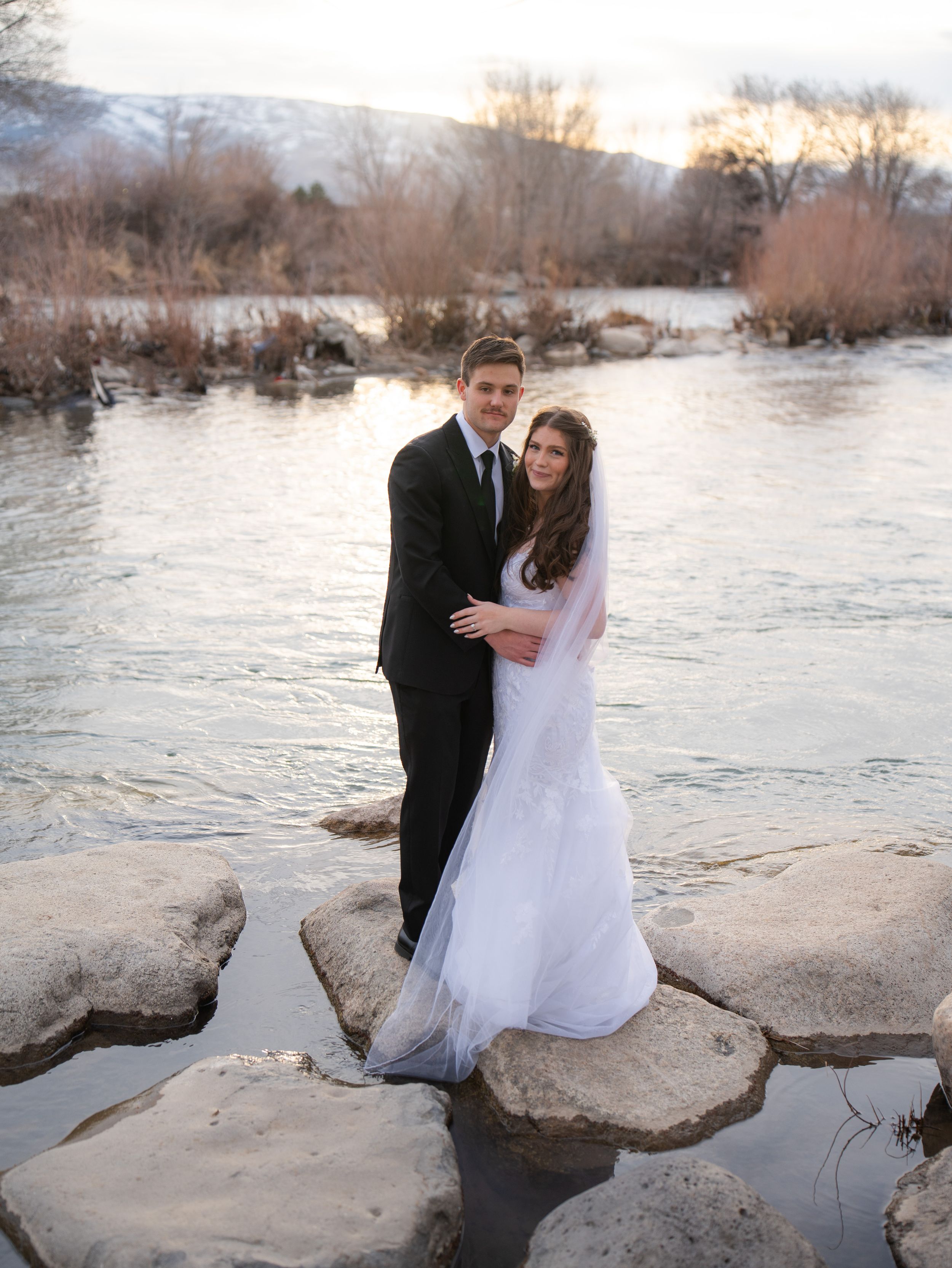 Couple in wedding attire sits together on large rocks by a serene river at sunset during a romantic photoshoot.