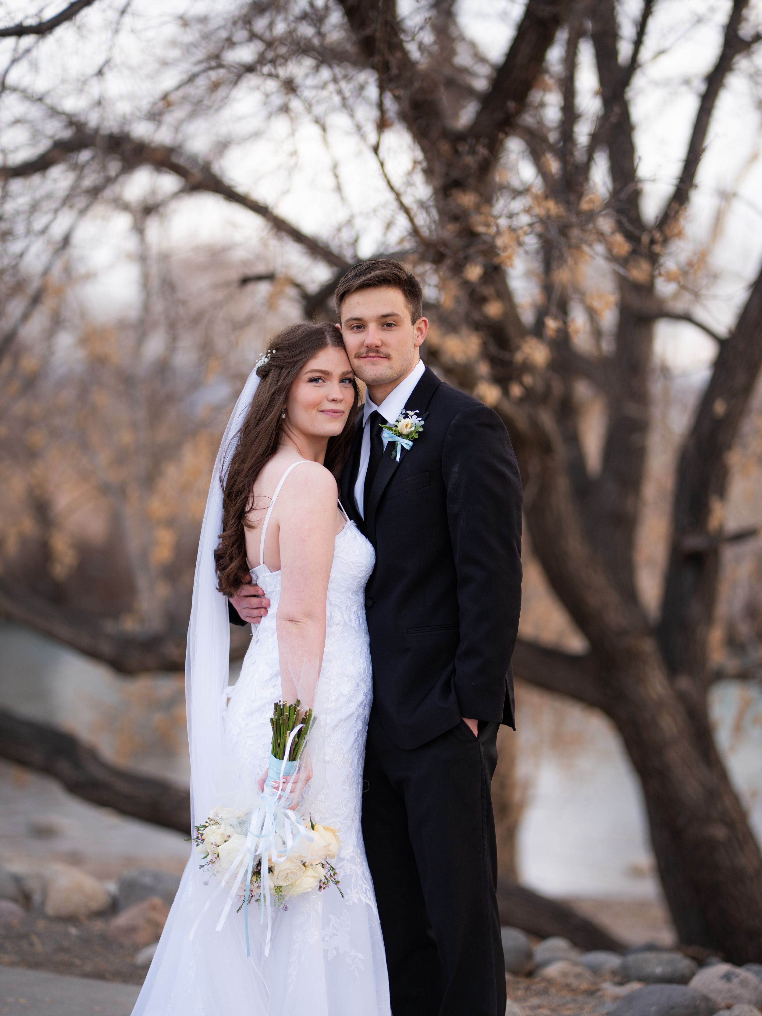 A romantic winter wedding photo shoot with a couple in formal attire embracing outdoors among bare trees.