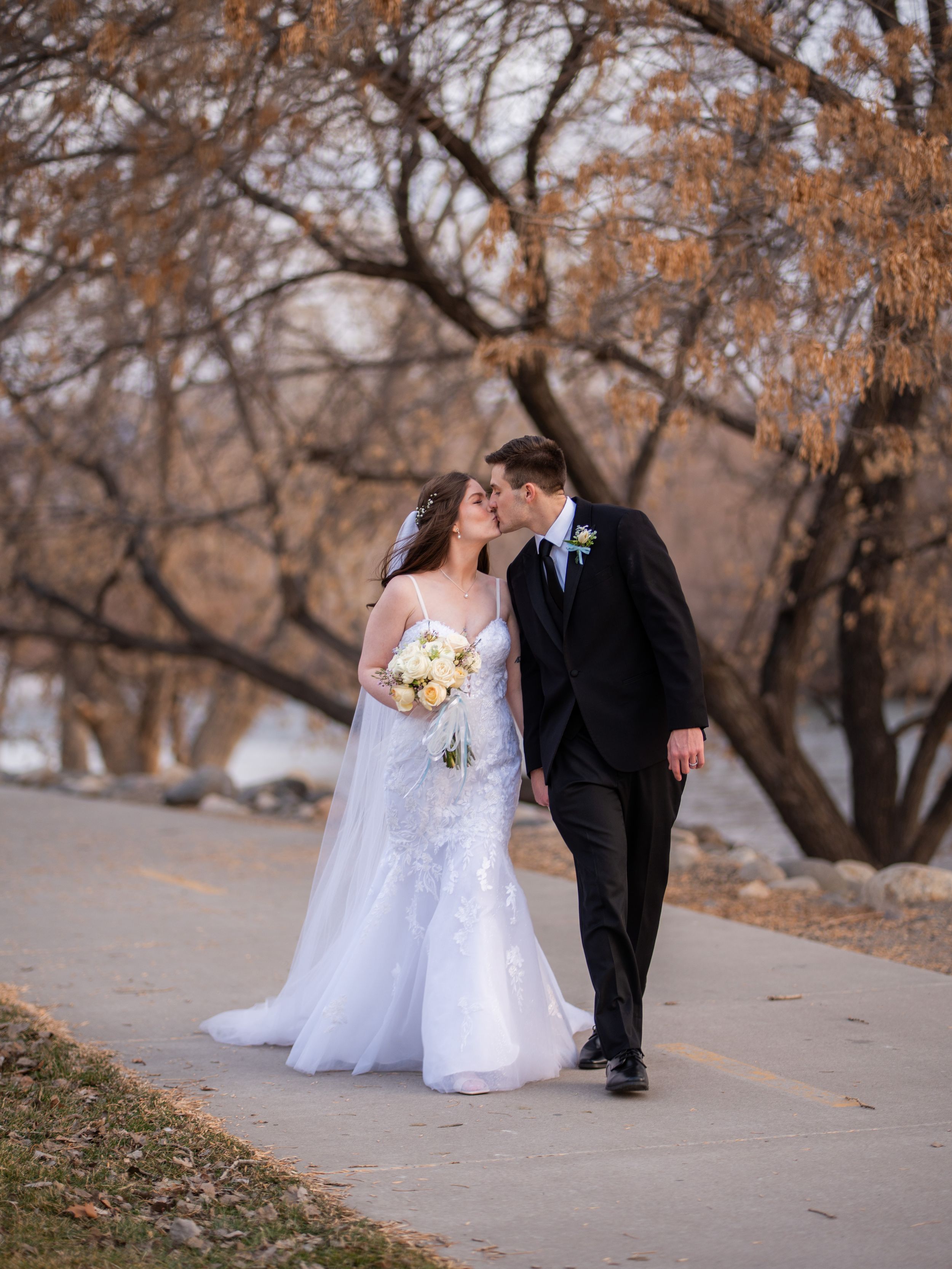 Newlywed couple shares romantic moments along a tree-lined path during autumn wedding photoshoot.