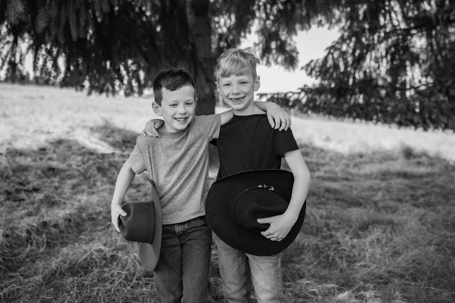 Two smiling boys hug outdoors in a black and white photo, one holding a cowboy hat.