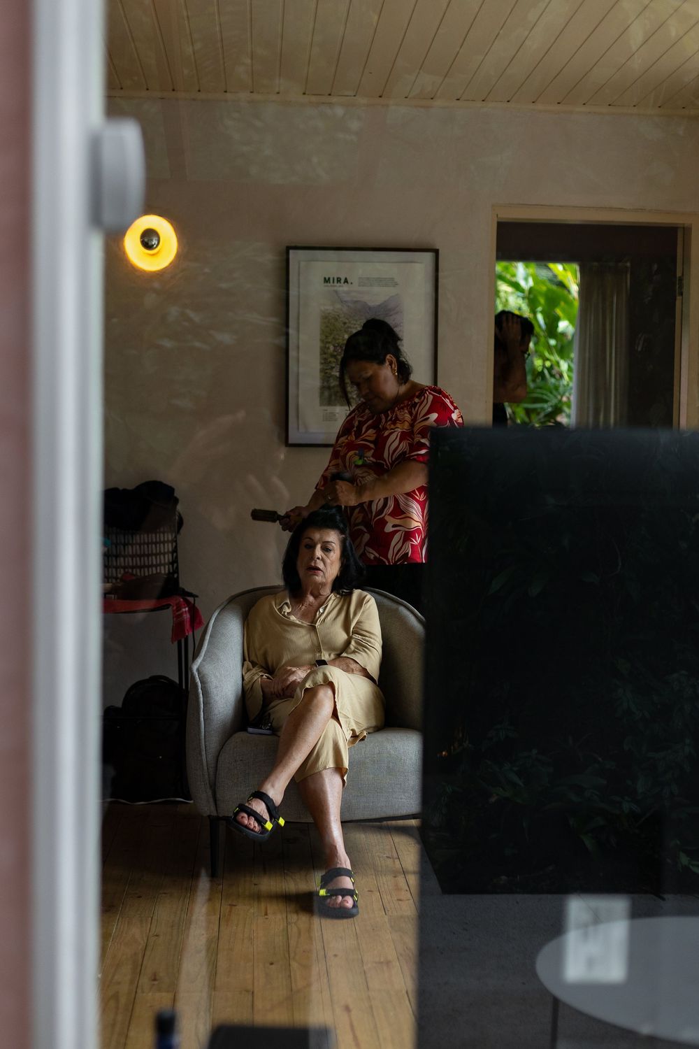 A person sits in a sunlit room with wooden floors and framed artwork on the walls, casting long shadows.