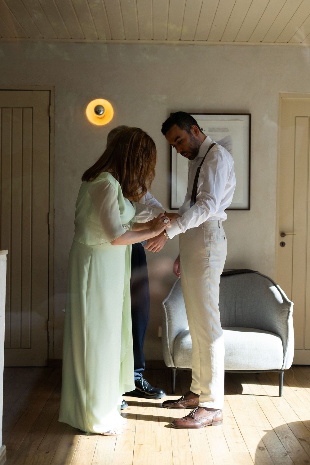 A healthcare worker in white uniform assists a patient in a green gown walking across a sunlit wooden floor.