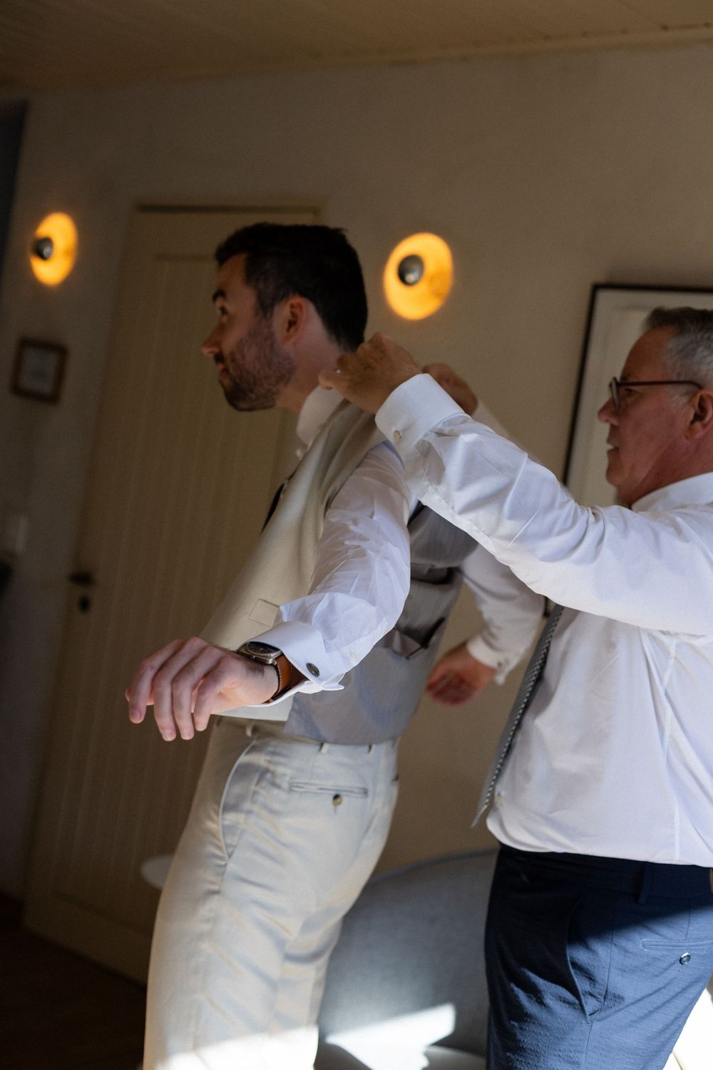 Two people in white shirts preparing formal attire in an interior room with warm lighting.