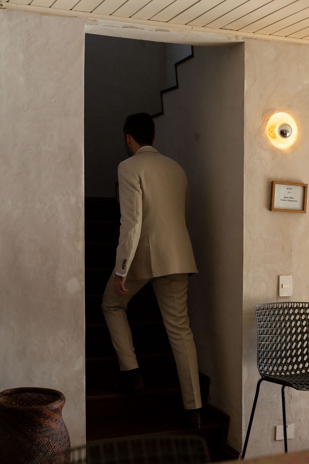 A person in a light-colored suit walks through a dimly lit hallway with wooden ceiling panels and wall sconces.