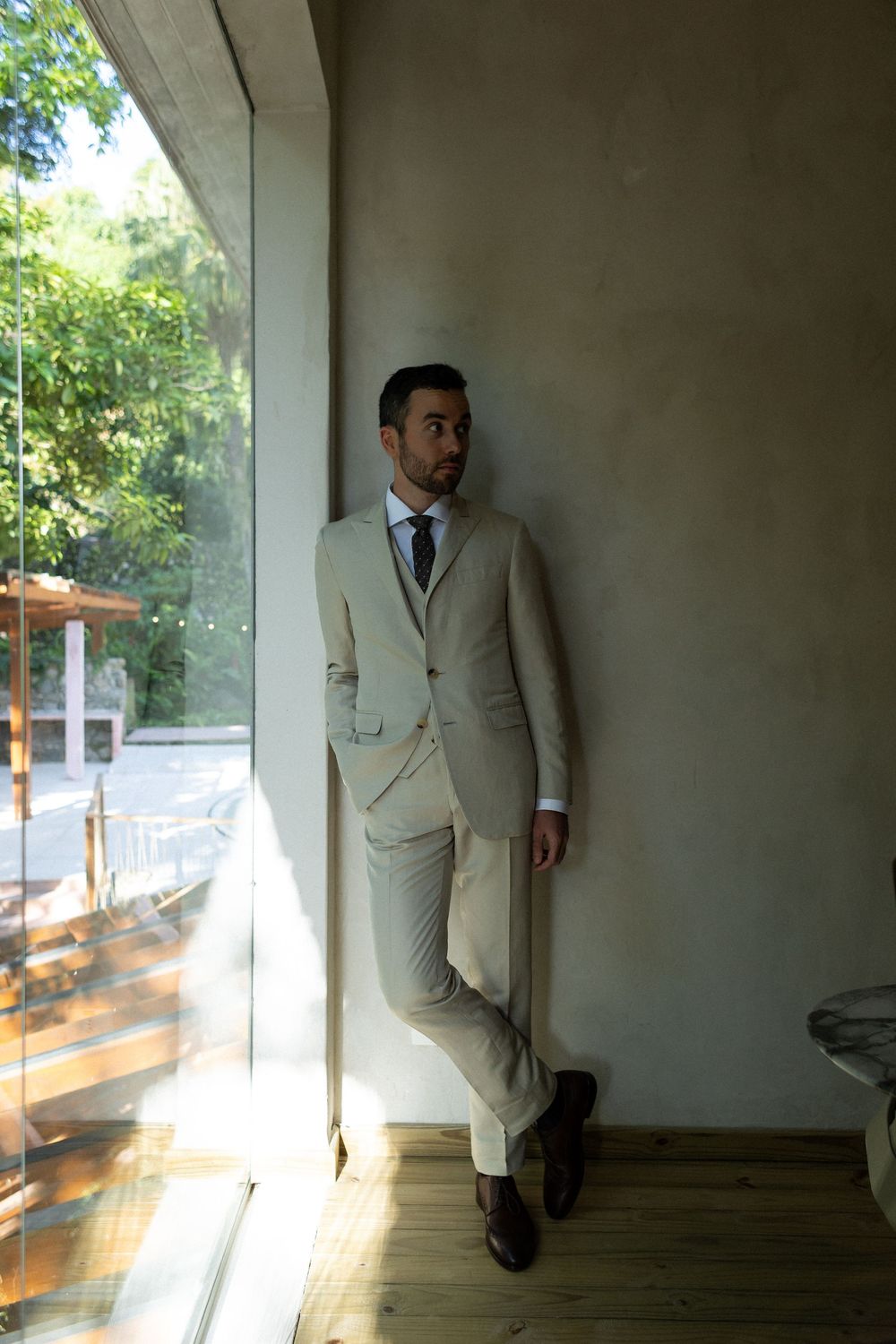 A figure in a light beige suit leans against a white wall in a sunlit hallway, creating a series of moody portraits.