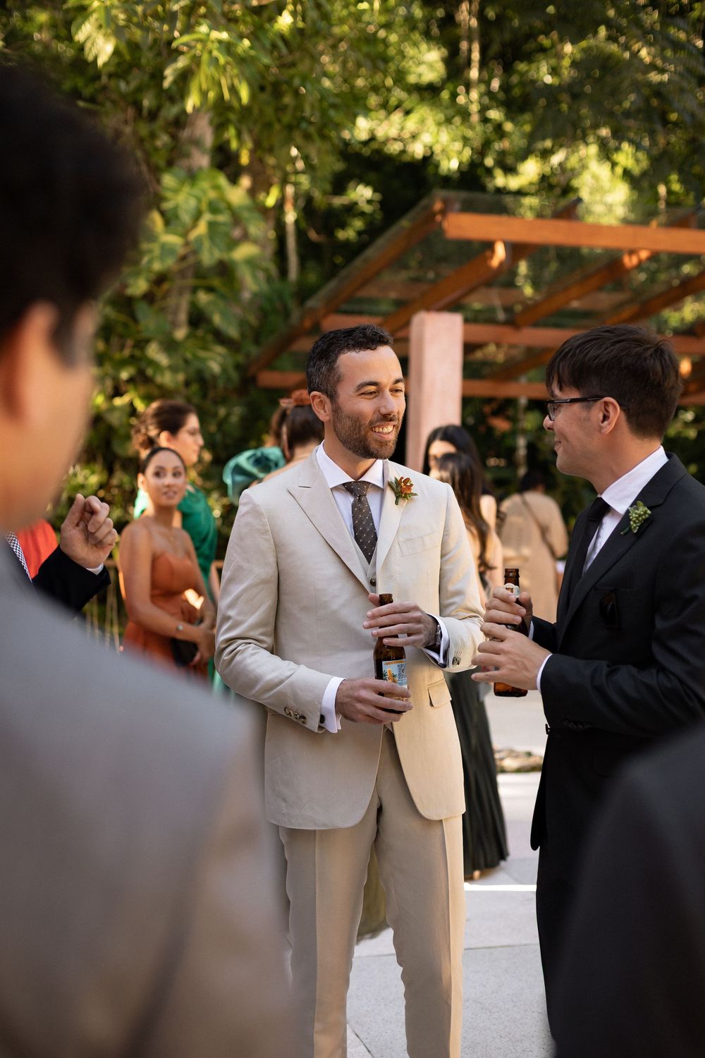 Wedding guests in formal attire gather outdoors under trees during a daytime celebration.