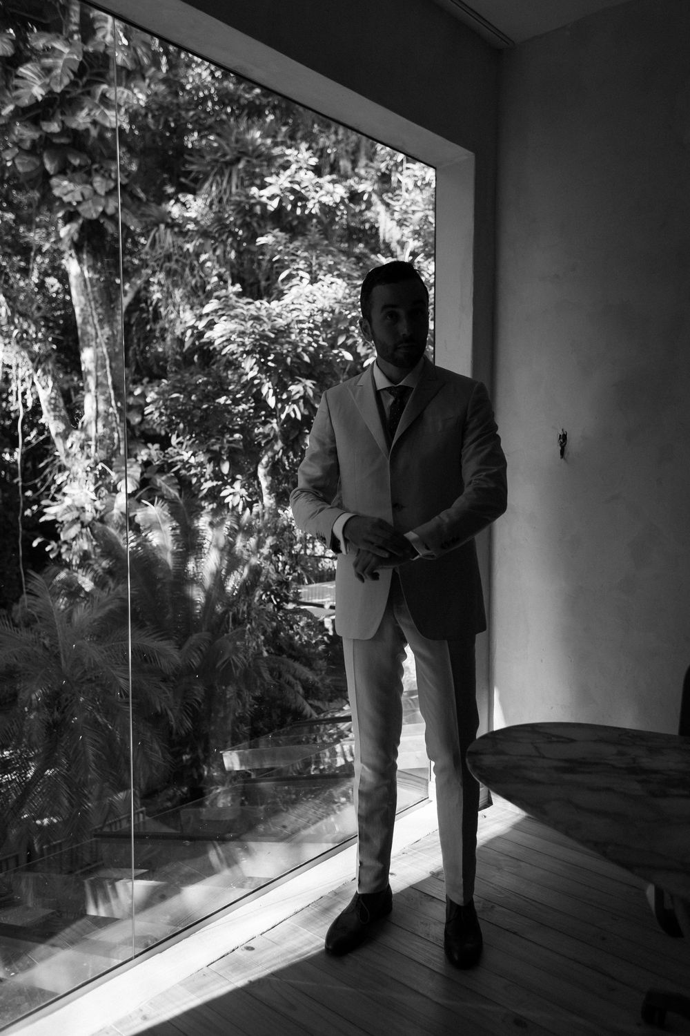 A person in a suit stands by large windows overlooking lush garden scenery in black and white.