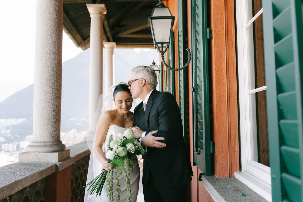 Bride and father share joyful moment on elegant columned terrace overlooking coastal view.