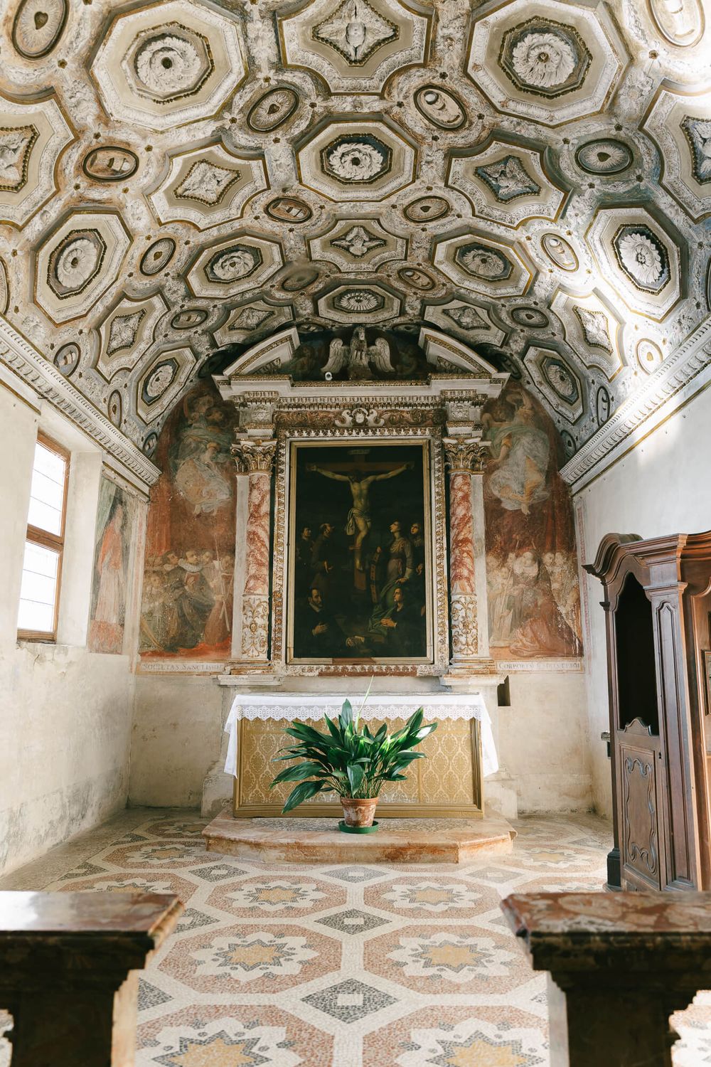 Historic chapel interior with ornate baroque ceiling, stone altar, and geometric floor patterns in muted earth tones.