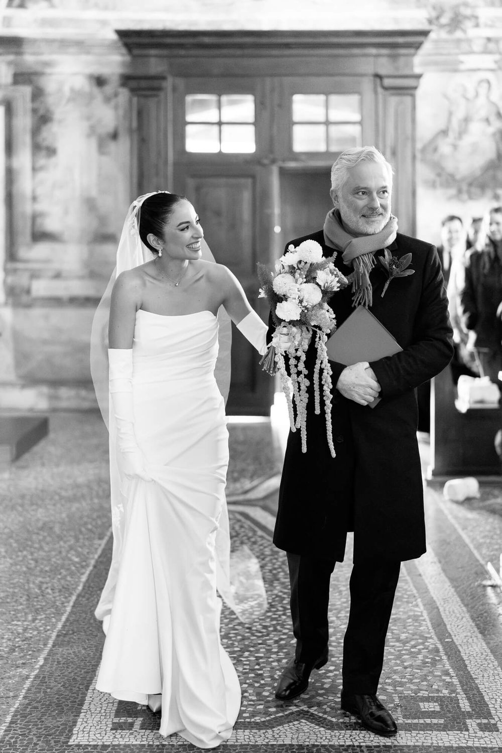 Black and white wedding photos show a bride in white dress walking arm in arm with an escort down a hallway.
