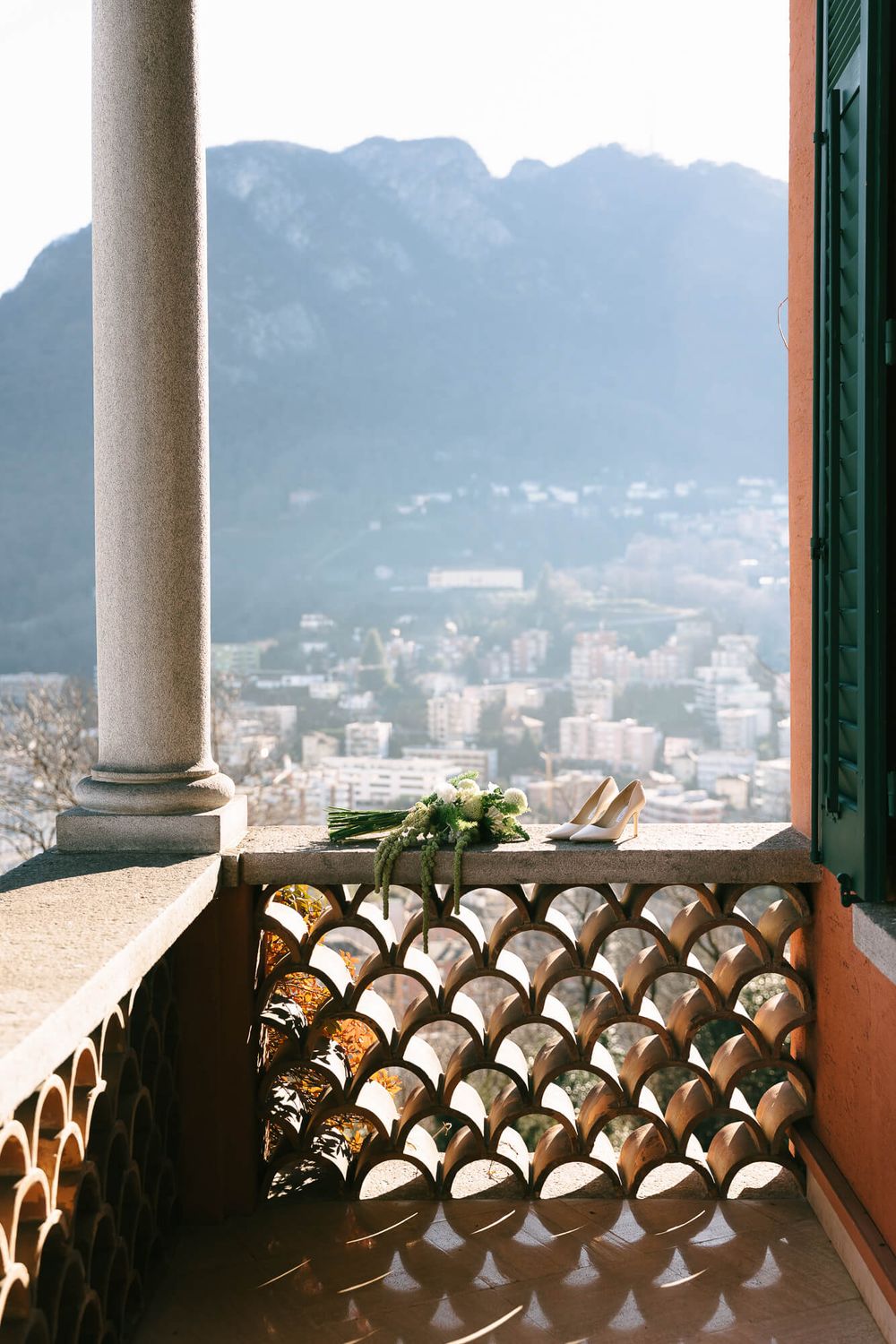 View of a mountain city through ornate balcony railing with a white column and green shutters in the foreground.