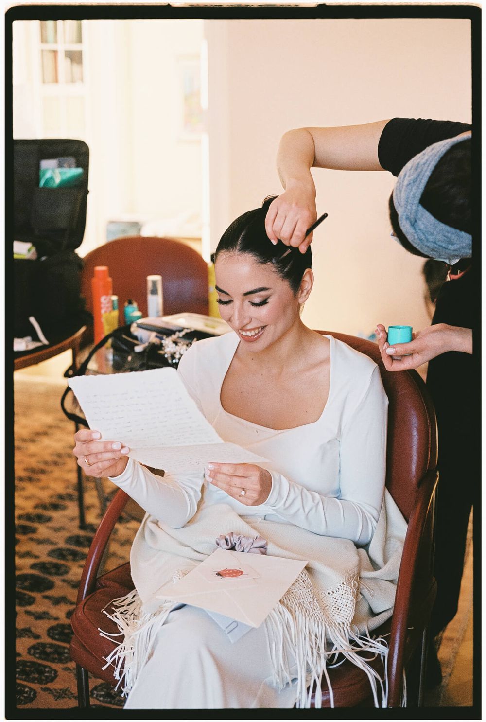 A bride in a white dress reads letters while getting ready on her wedding day in a warmly lit room.
