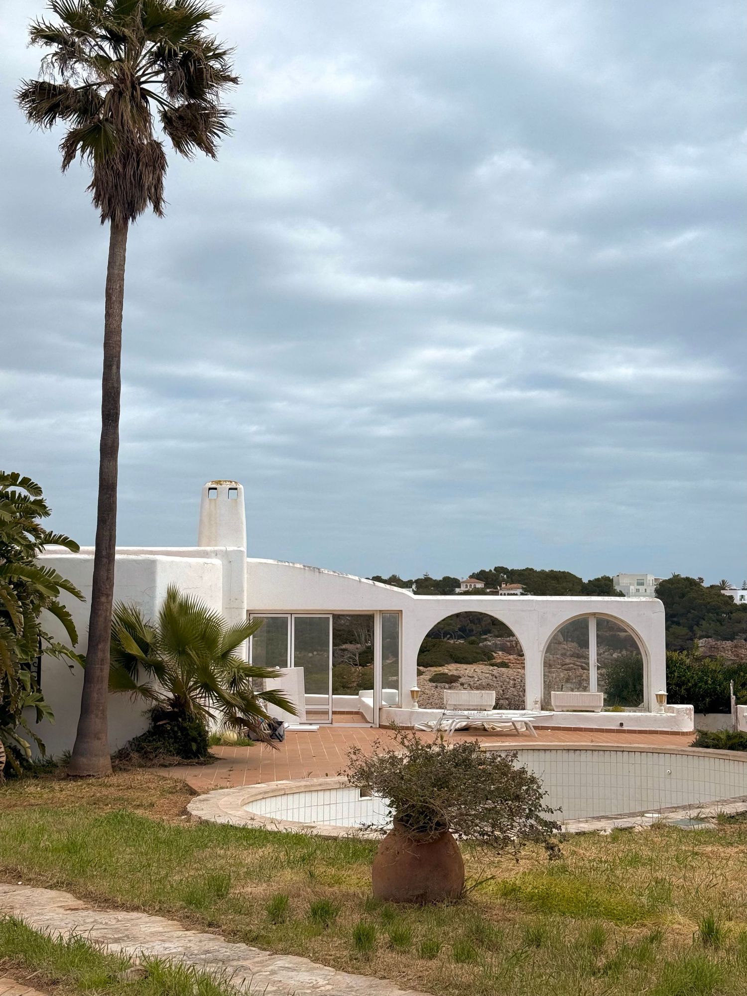 A modern, white building with arched entryways surrounded by palm trees and greenery against a cloudy sky.
