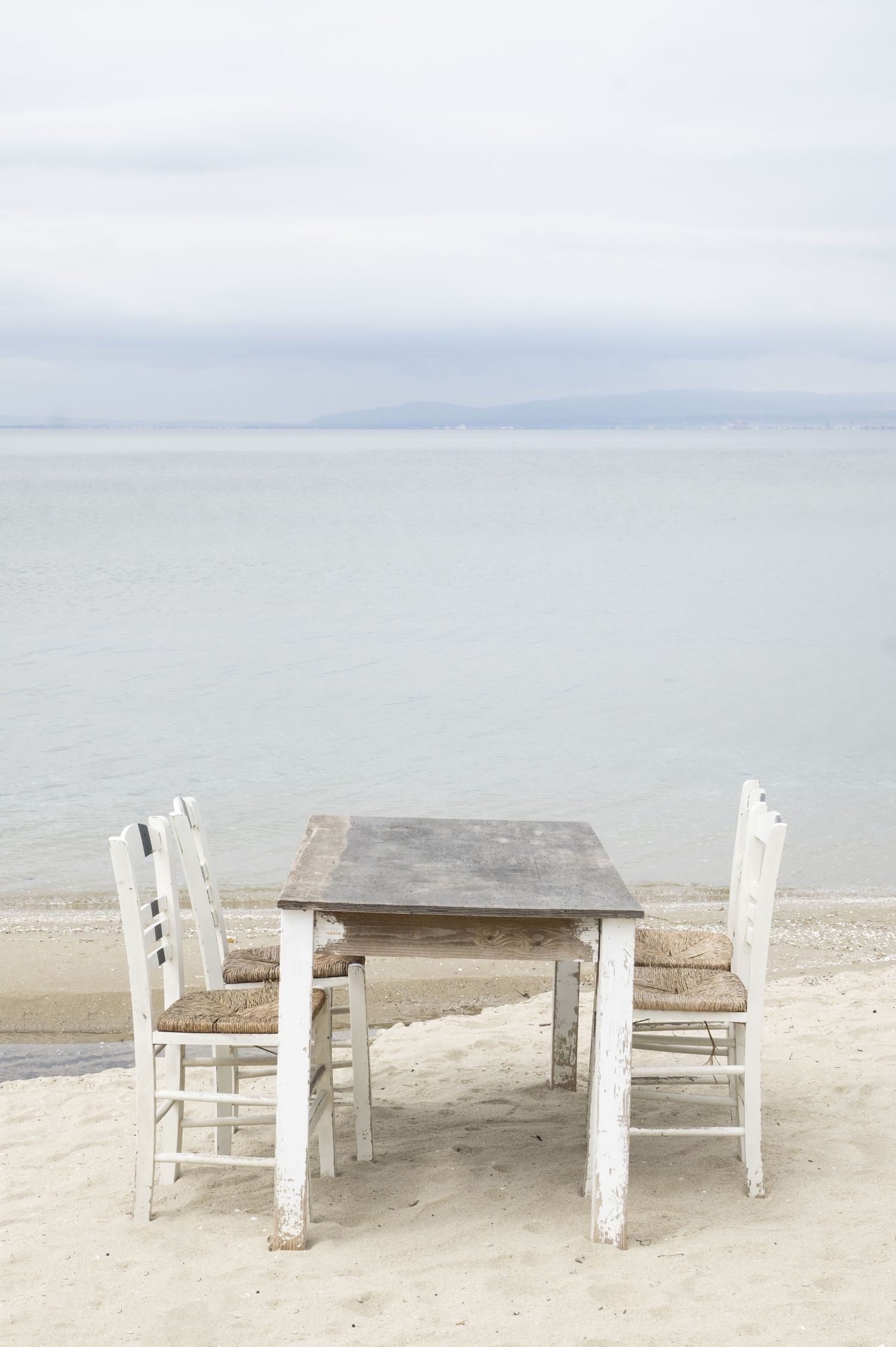 Weathered white wooden picnic tables and benches sit empty on a sandy beach against a misty ocean backdrop.