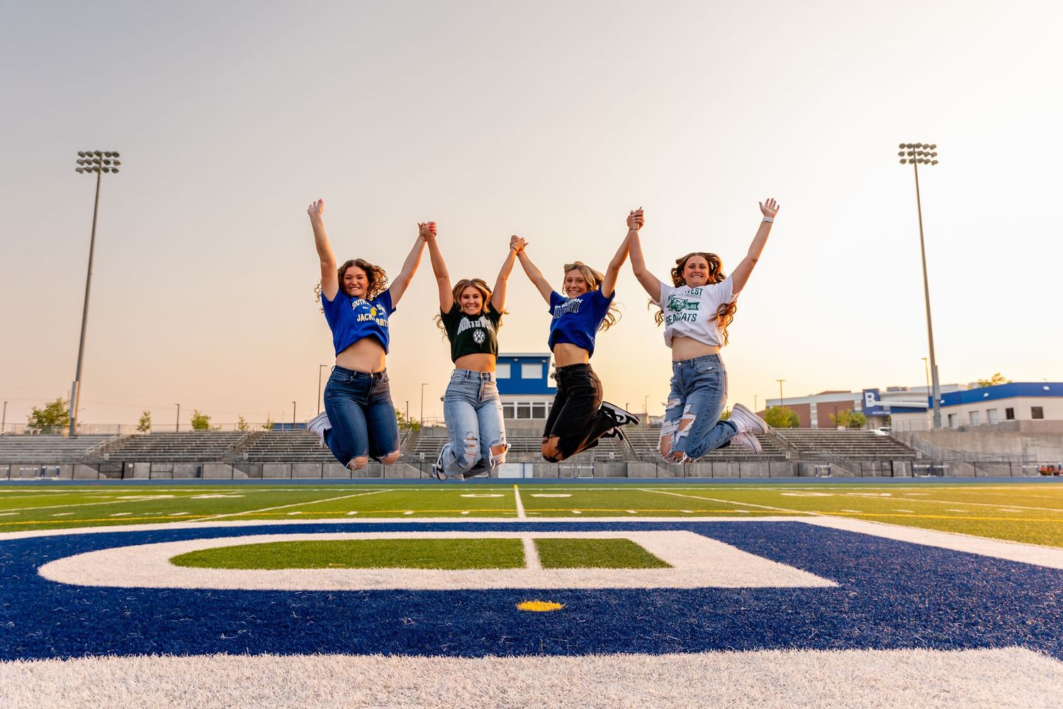 Cheerful high school graduates, wearing college tshirts & jumping in Bennington High School stadium with arms raised in celebration.