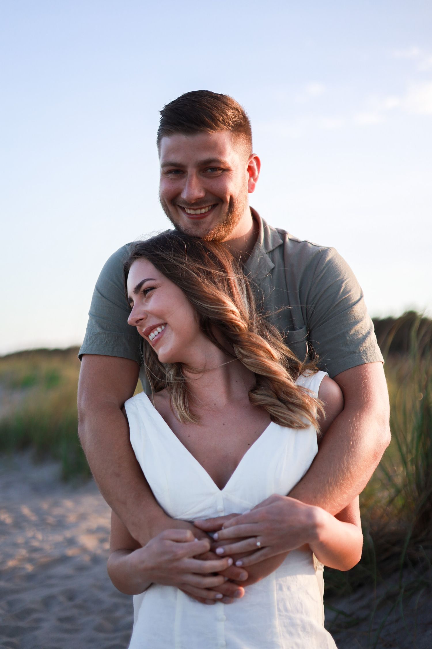 Happy couple embracing on a sandy beach at sunset, woman in white dress laughing joyfully.