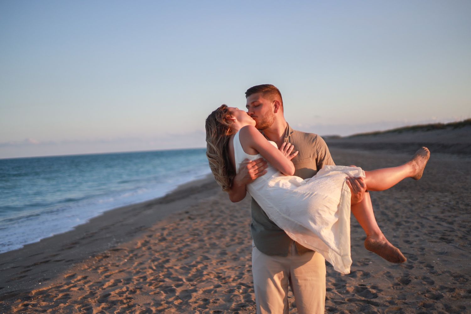 Romantic couple sharing a kiss on the beach at sunset, man lifting woman in white dress.