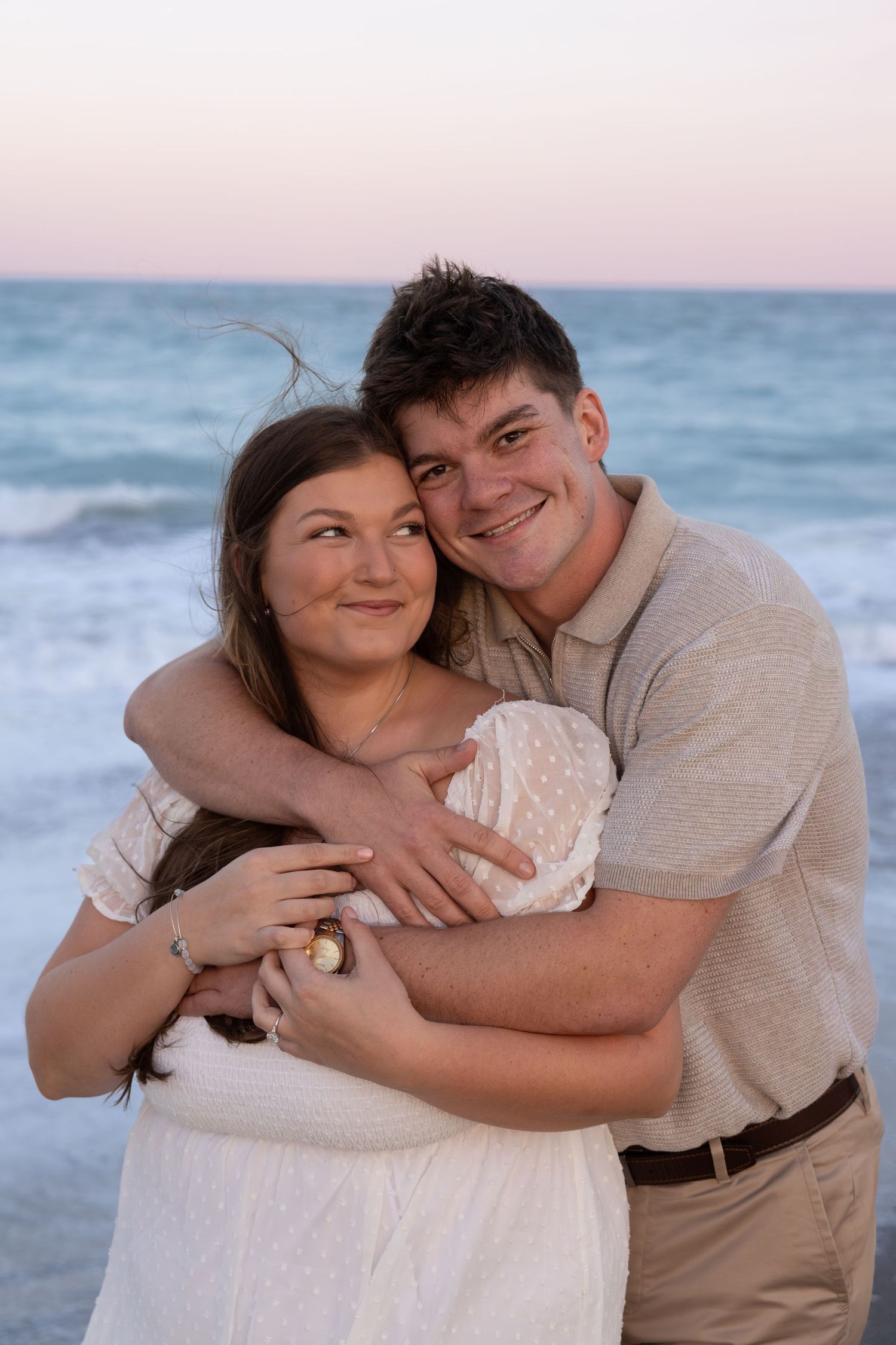 Happy young couple embraces warmly on beach with soft pastel ocean sunset in background.