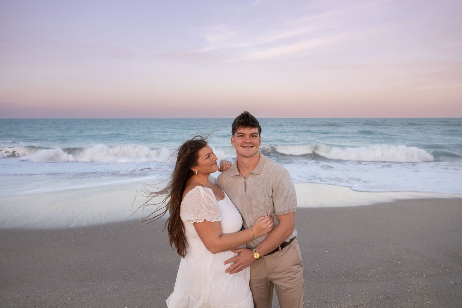 Smiling couple poses together on sandy beach shoreline with pastel pink ocean sunset backdrop.