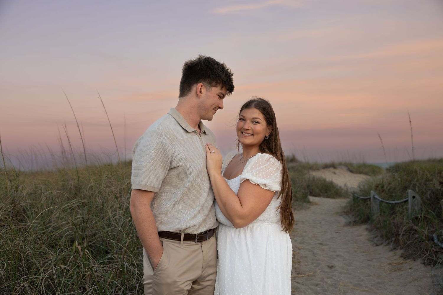 Young couple smiles together on a sandy beach path with sea oats and pastel sunset sky.