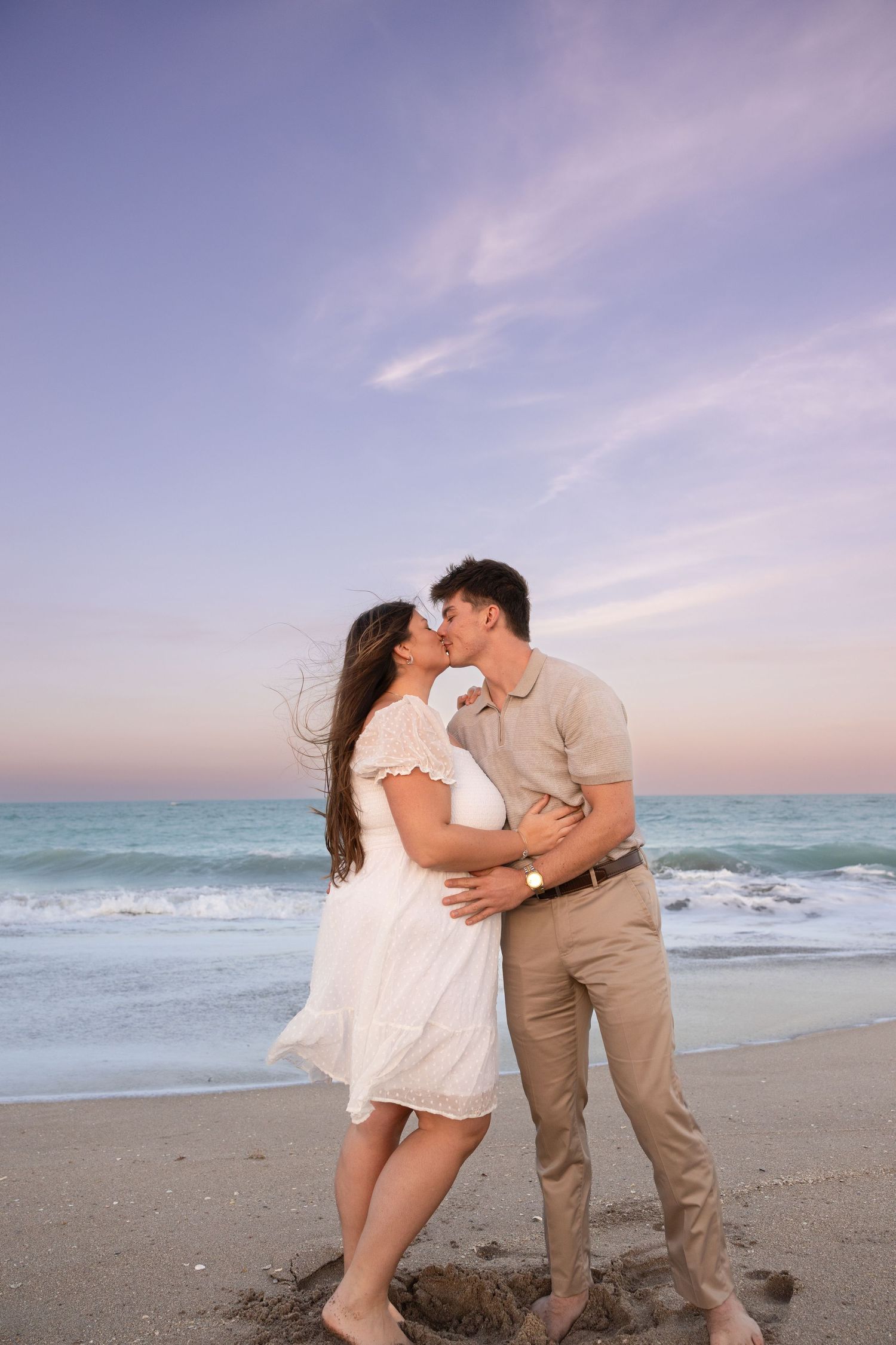 Couple shares romantic kiss on beach shoreline with turquoise ocean waves and purple sunset sky.