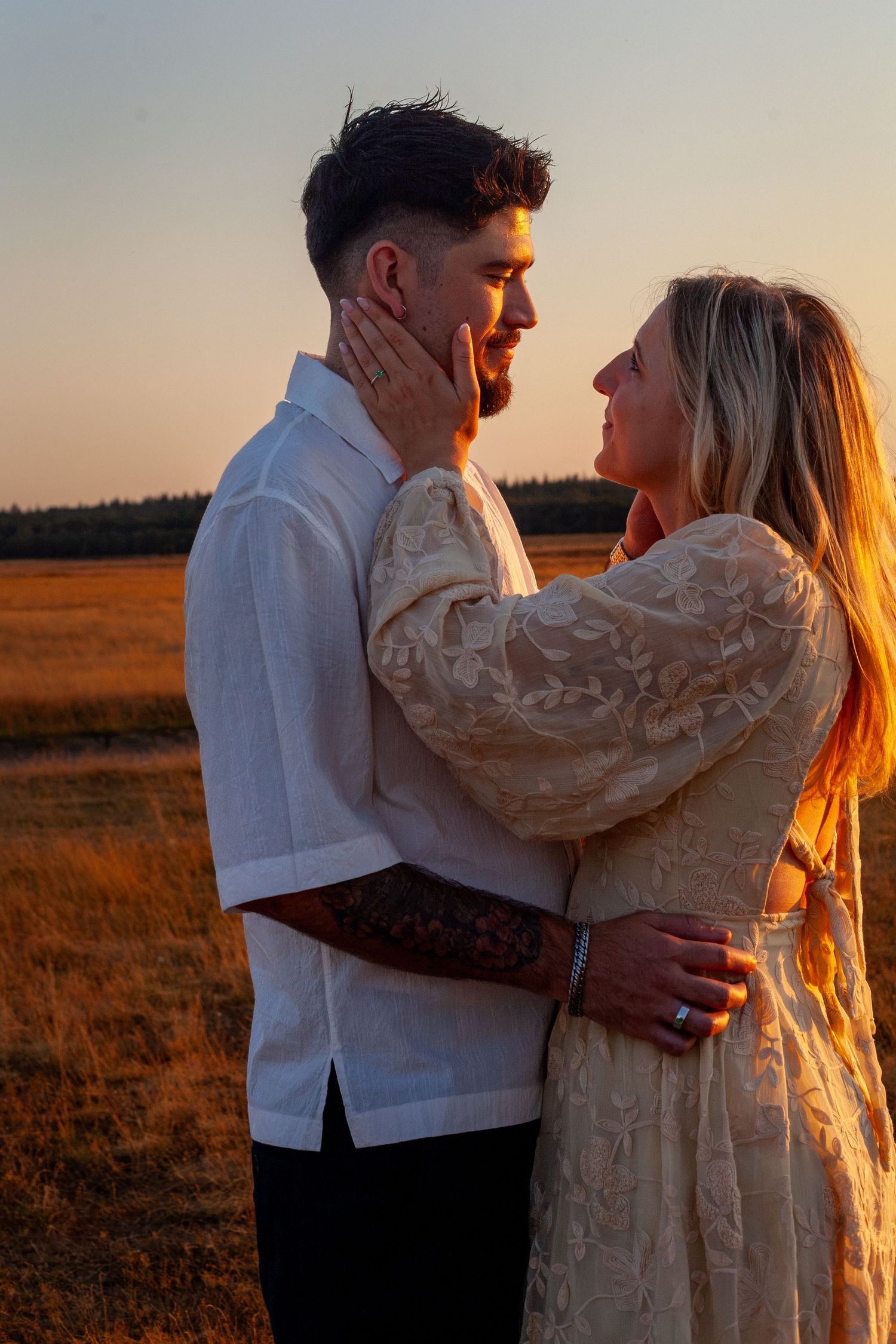 Couple embracing at golden hour sunset in a field, woman in lace dress touching mans face romantically.