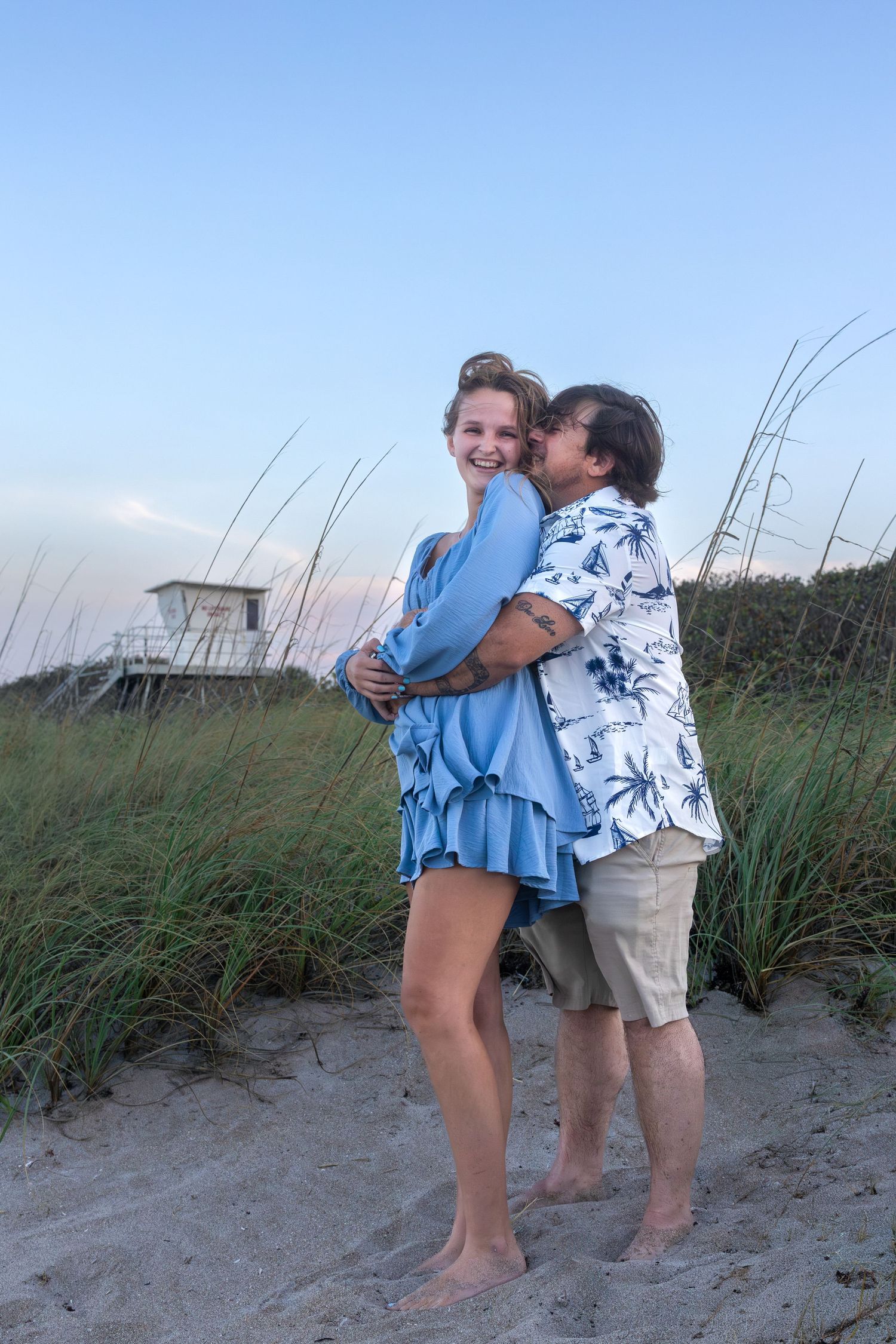 Couple embracing and kissing on sandy beach dunes at twilight near sea grass.