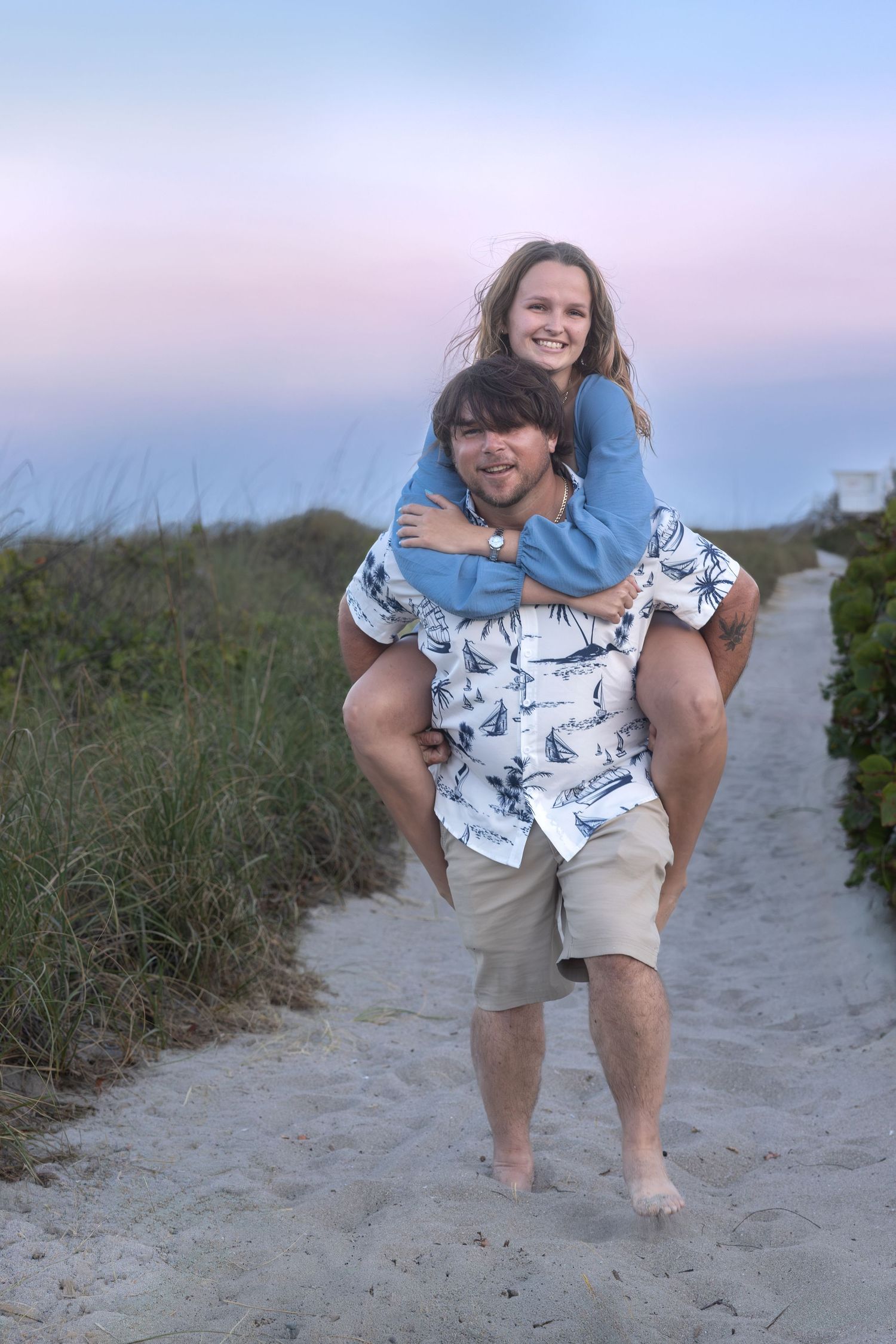 Man giving woman piggyback ride on sandy beach path at dusk with pastel sky.