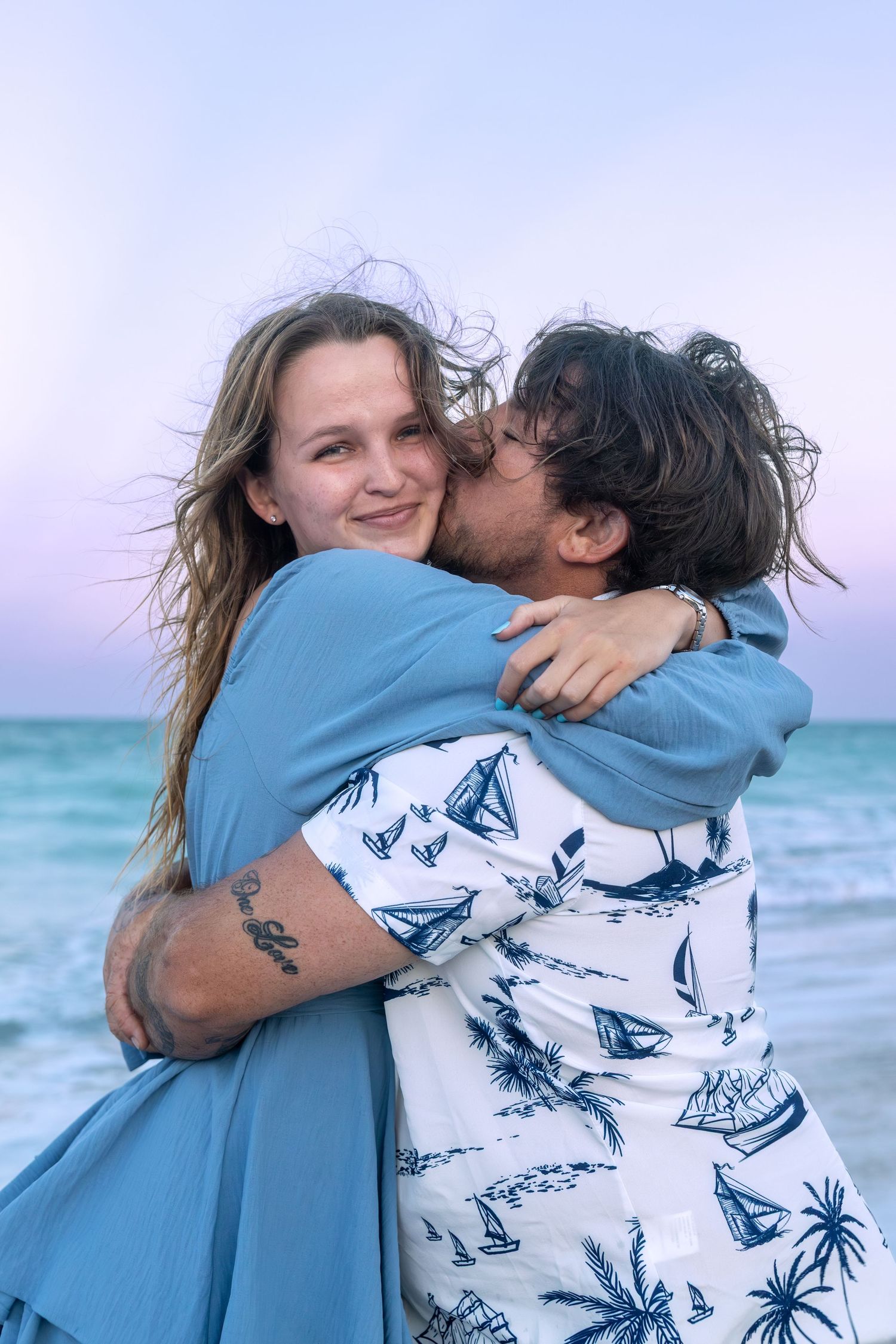 Couple hugging and kissing on beach at sunset with turquoise ocean in background.