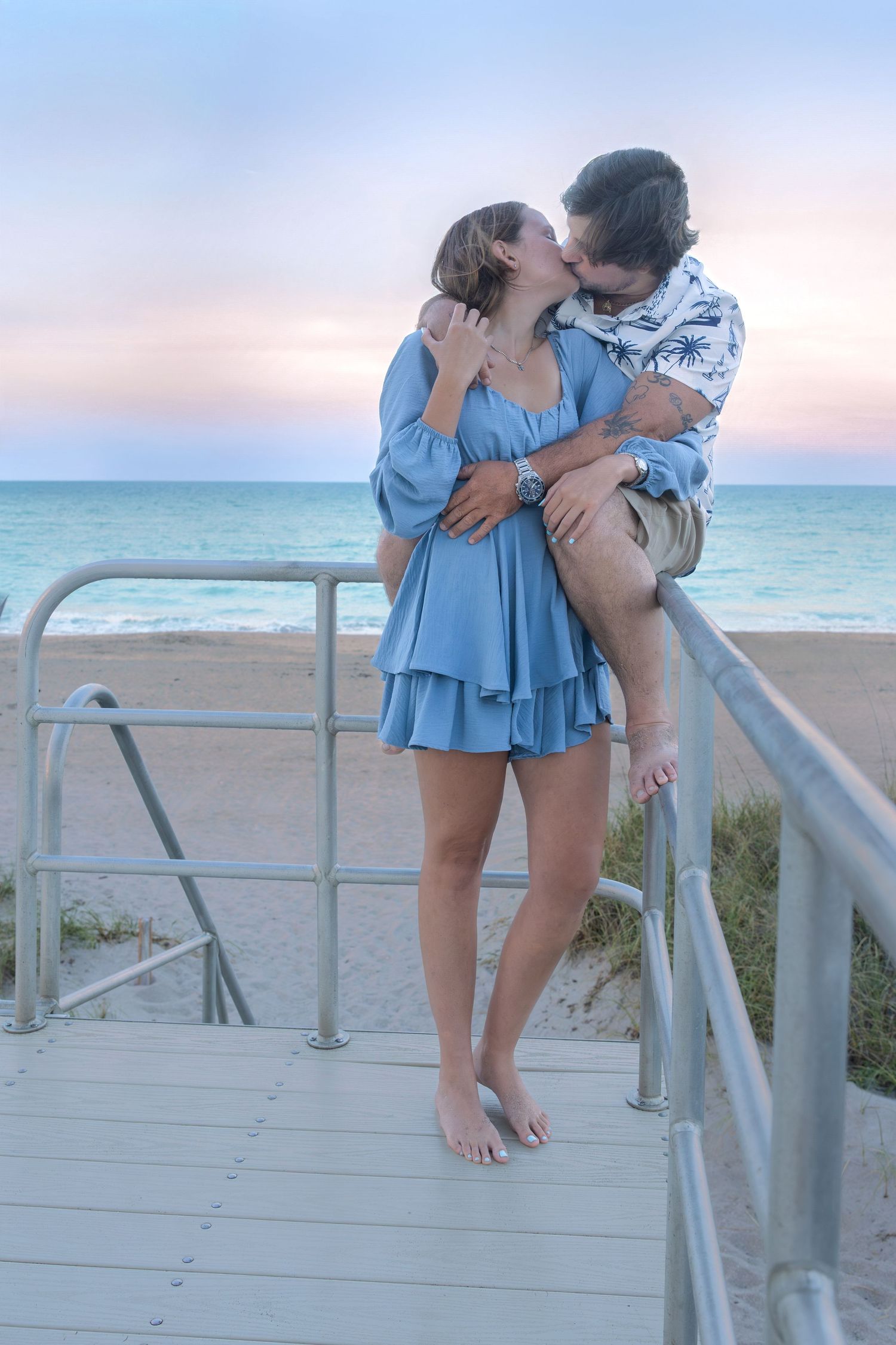 Couple sharing romantic kiss on wooden beach boardwalk railing at colorful sunset.