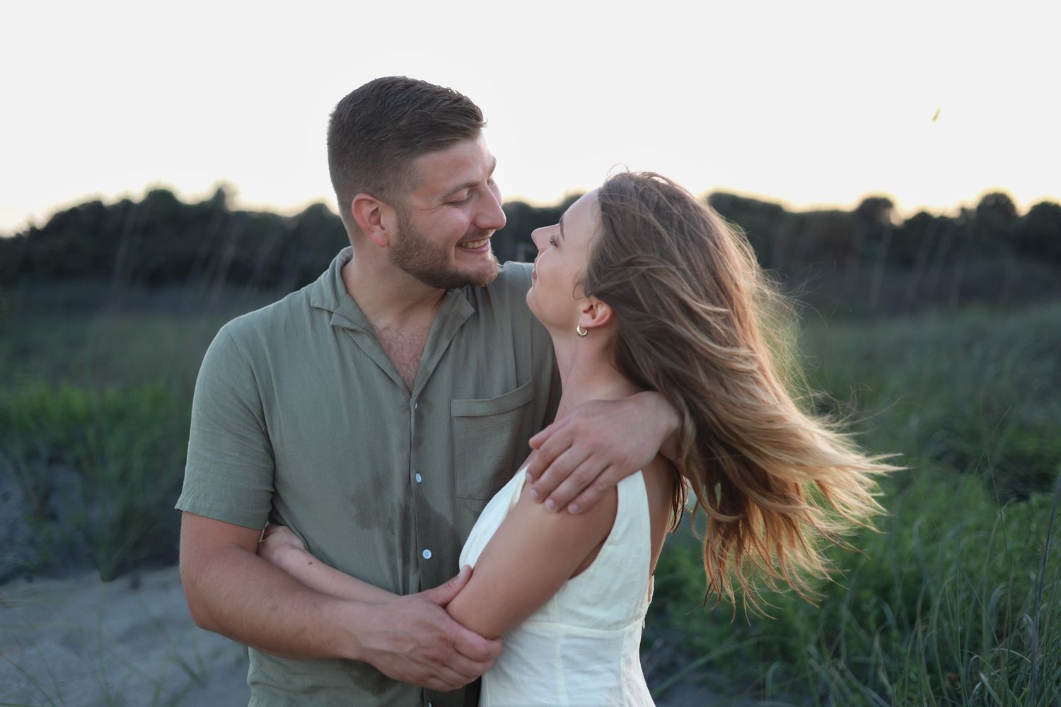 Joyful couple laughing together on a beach at golden hour with green dunes behind them.