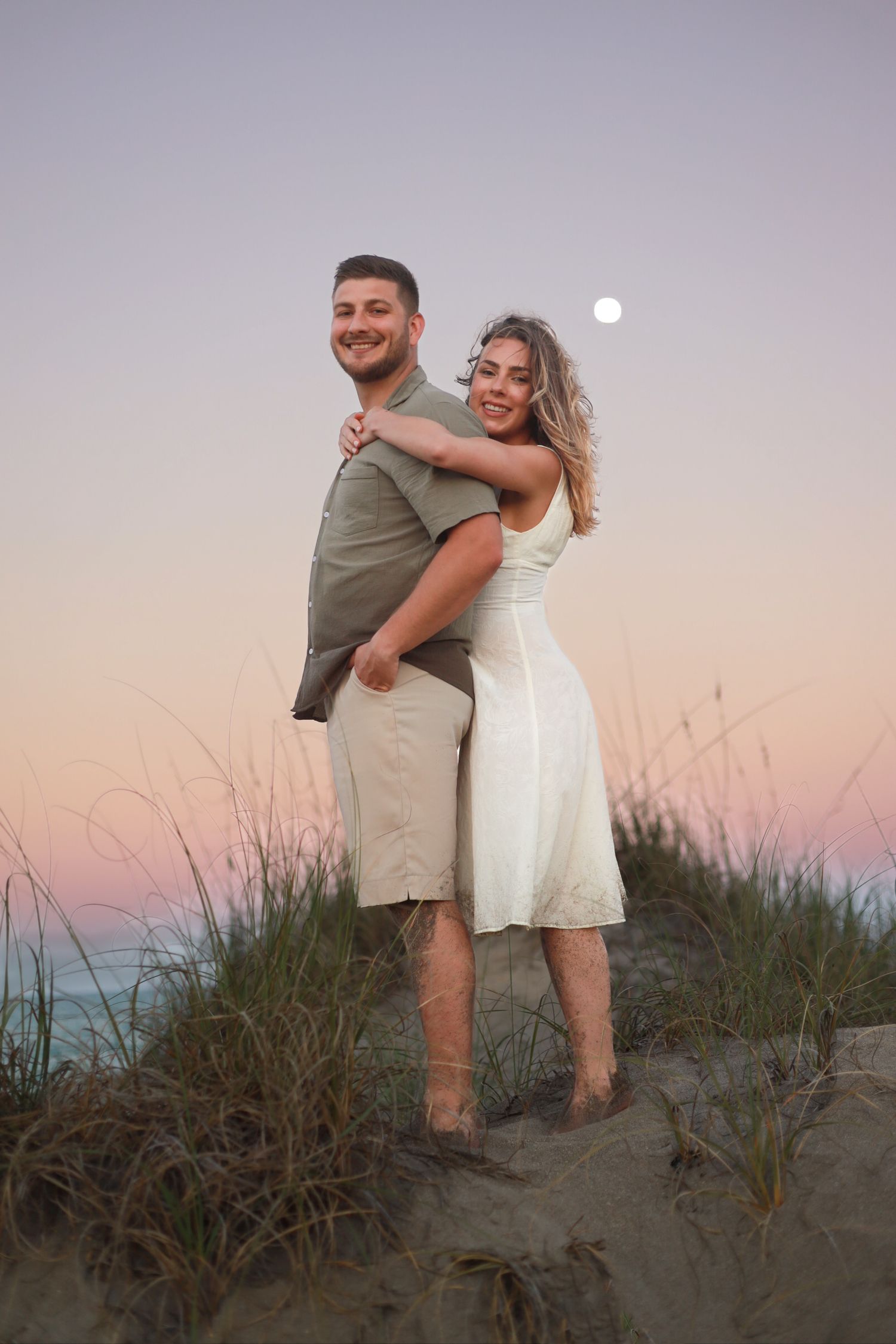 Smiling couple posing on a sand dune at sunset with a full moon visible in the sky.