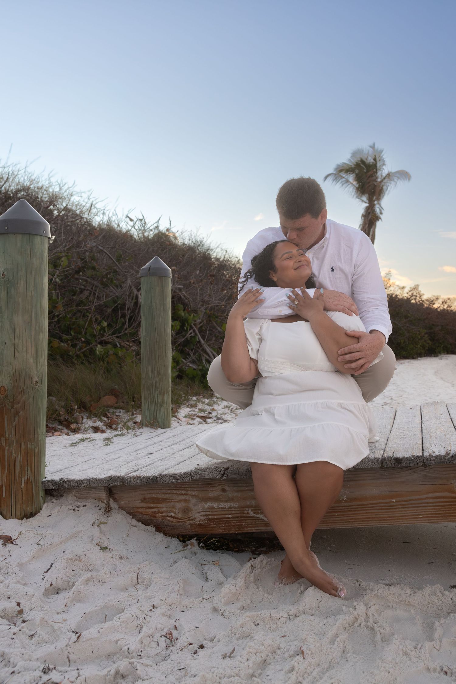 Couple sharing tender moment sitting on wooden beach boardwalk at dusk.