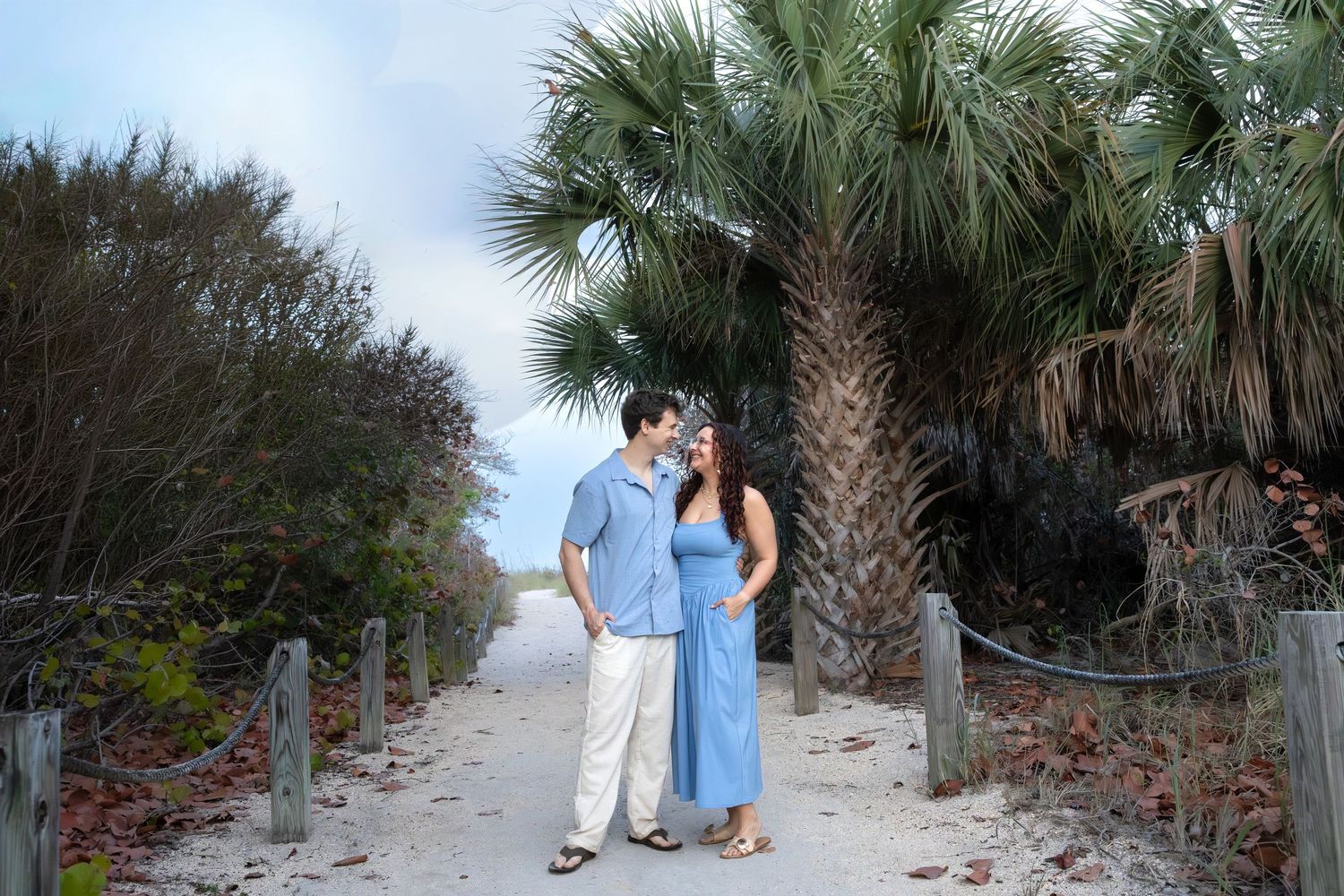 A couple shares a romantic moment on a sandy path flanked by palm trees at a coastal beach location.