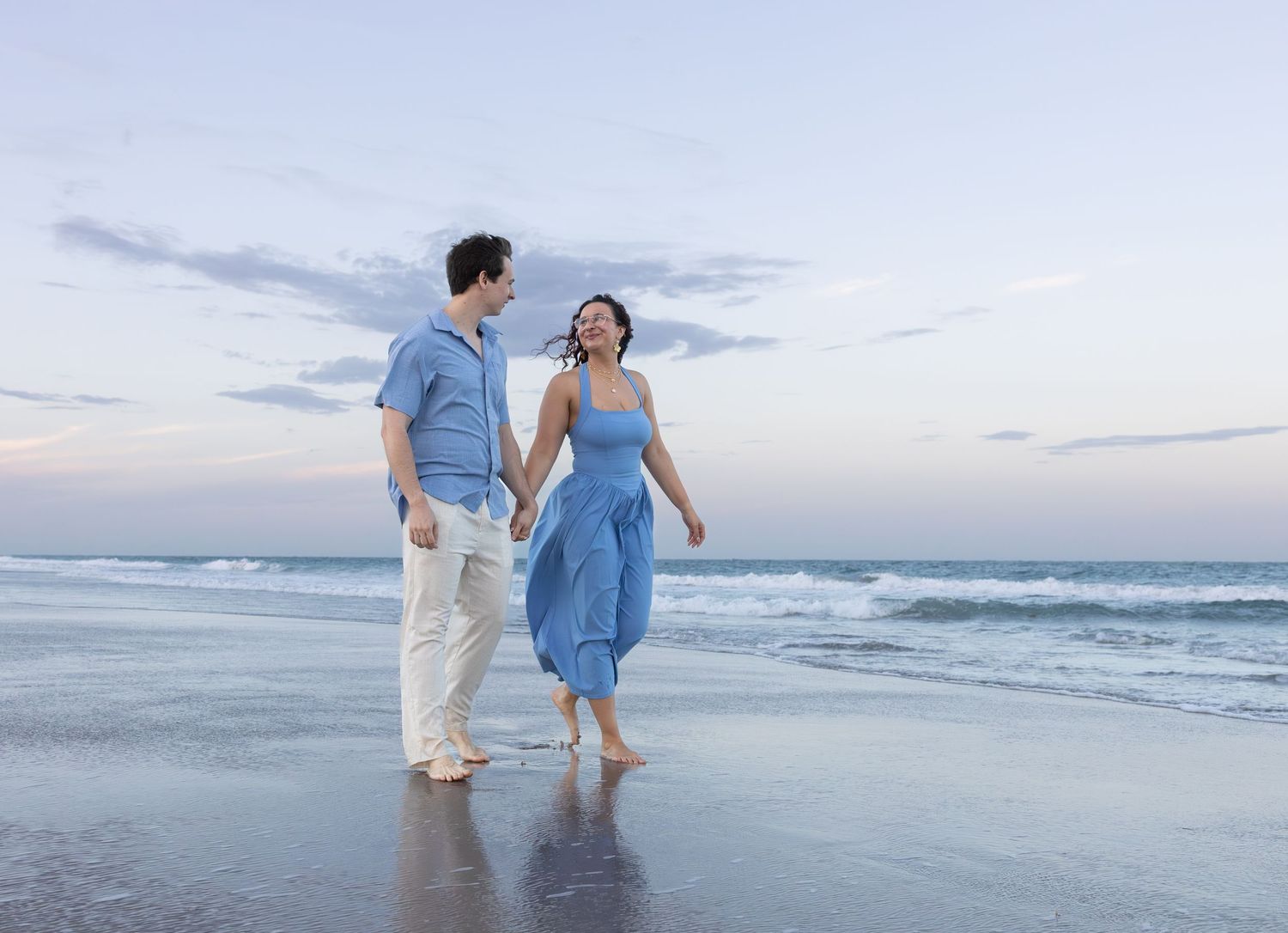 A happy couple walks hand-in-hand along a wet sandy beach at dusk with calm ocean waves behind them.