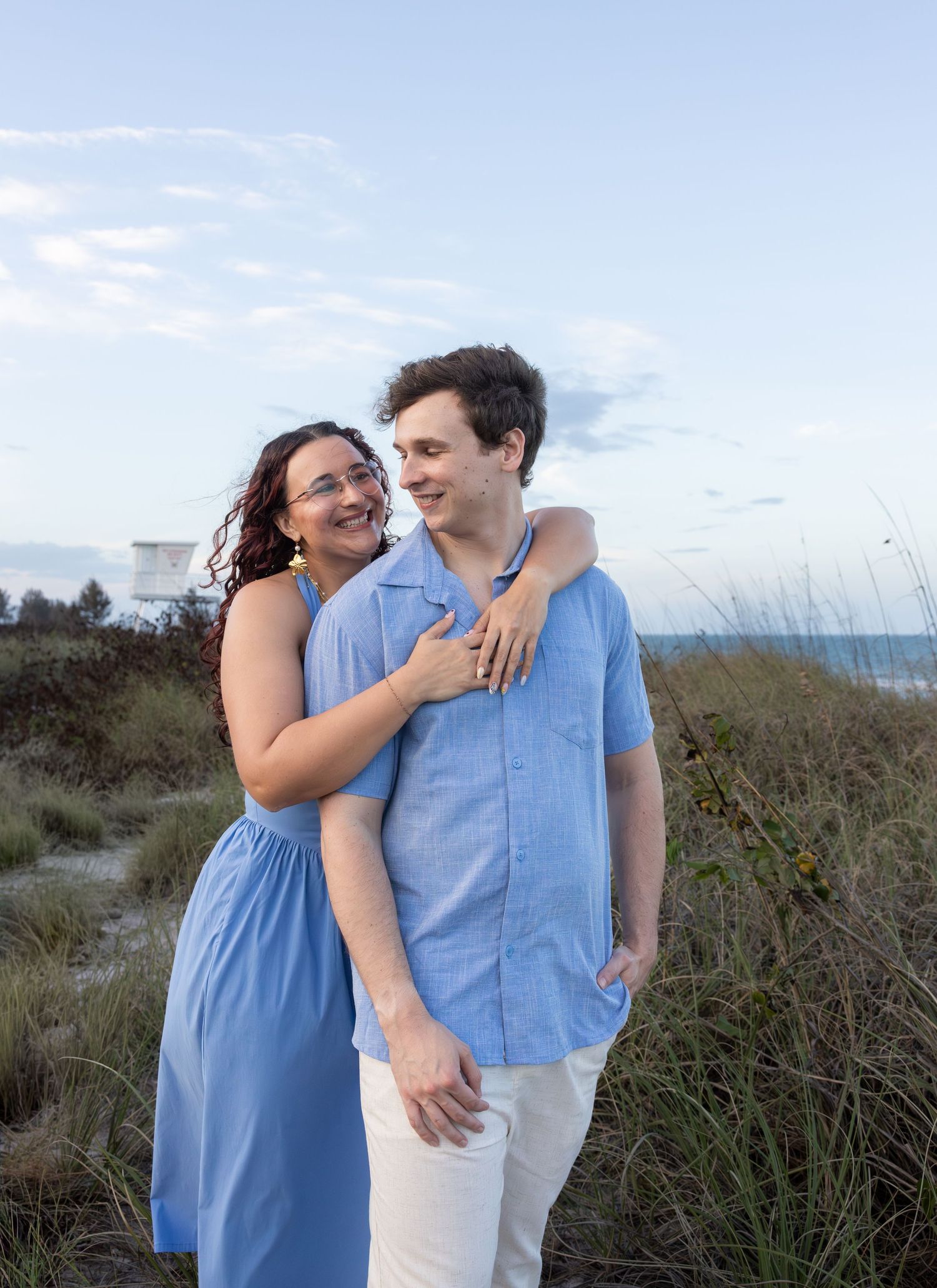 A smiling woman embraces a young man on a sandy beach path surrounded by sea grass at twilight.