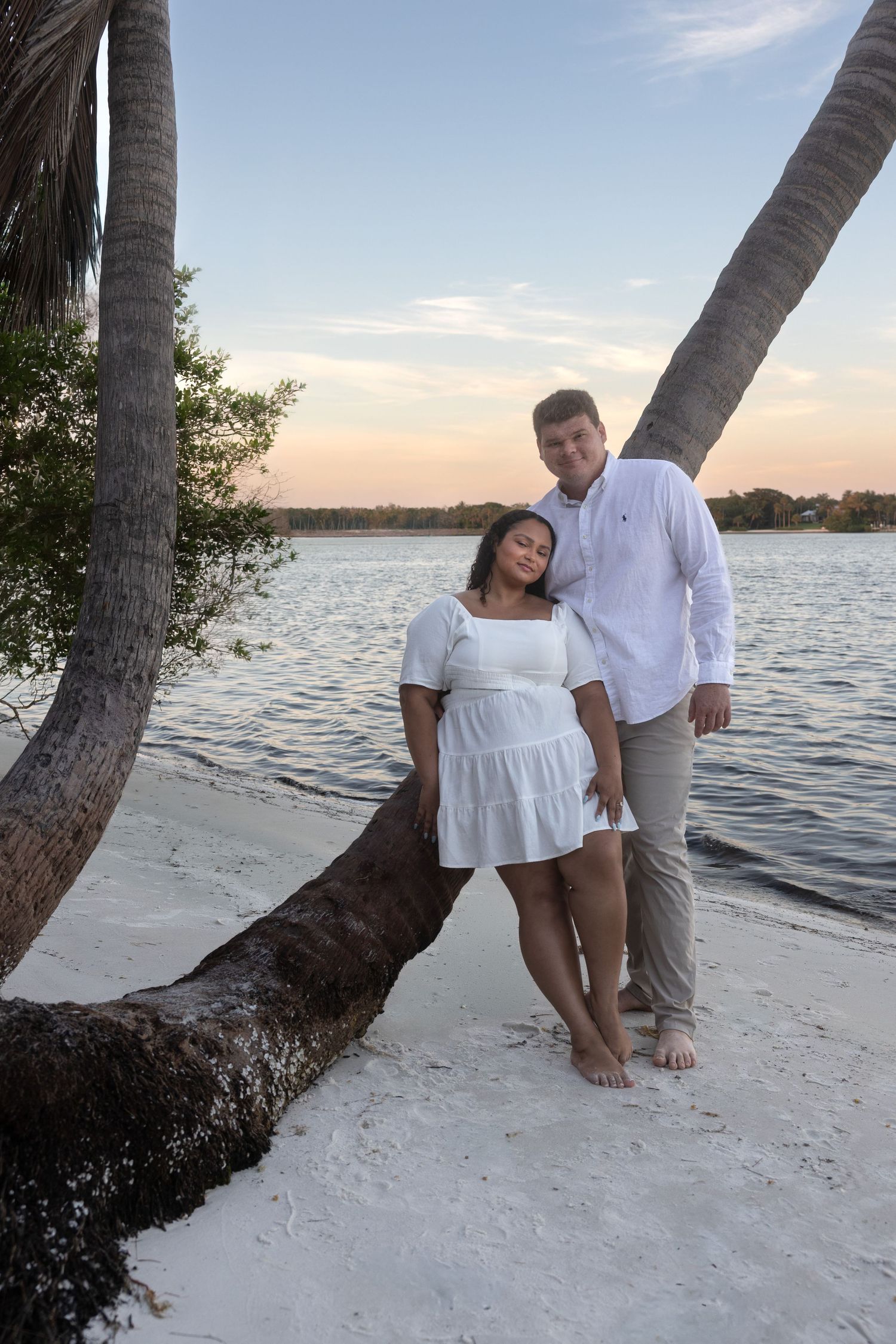 Couple in white outfits posing under leaning palm trees on sandy beach at sunset.