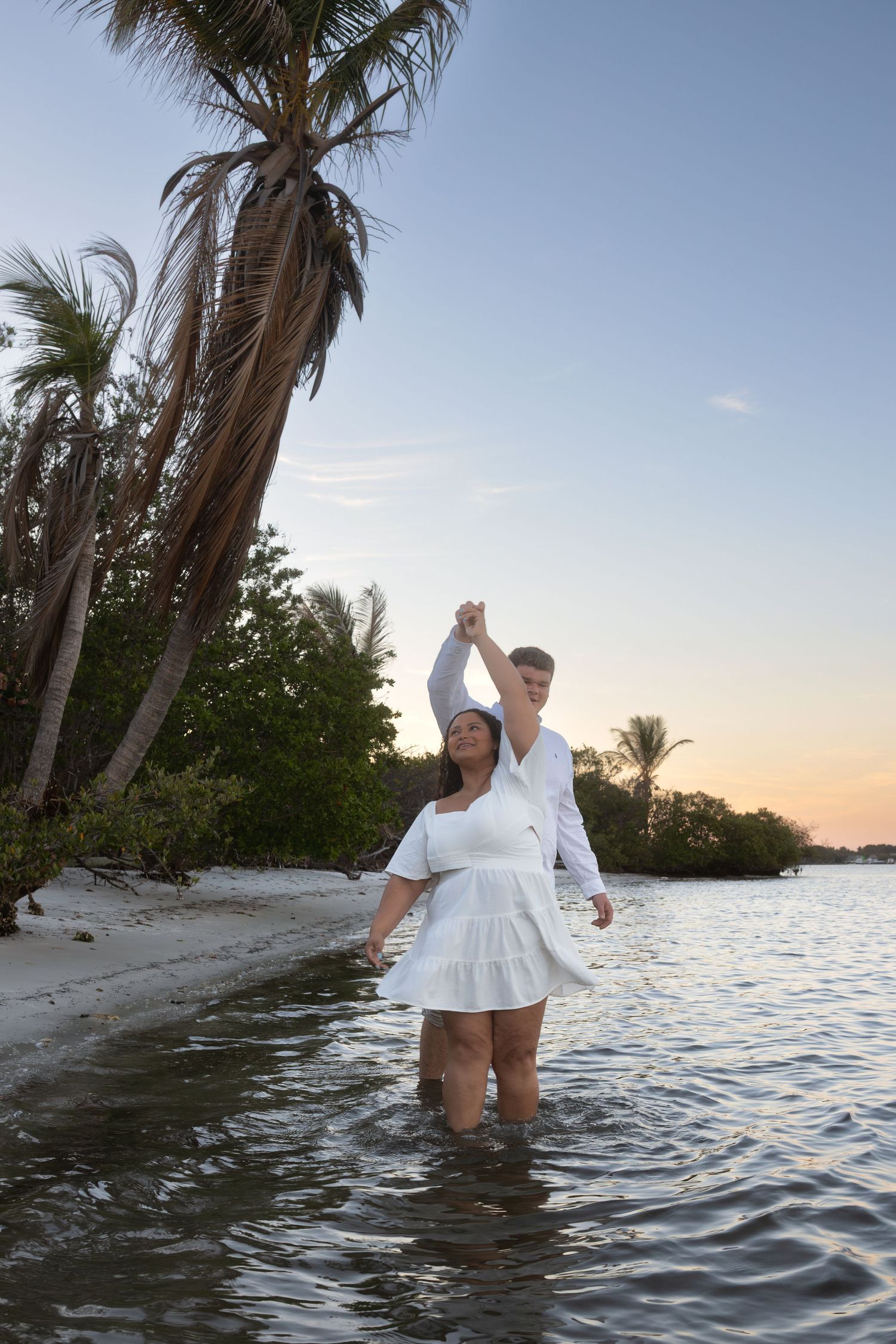 Couple in white outfits dancing on a tropical beach at sunset, wading in shallow water near palm trees.