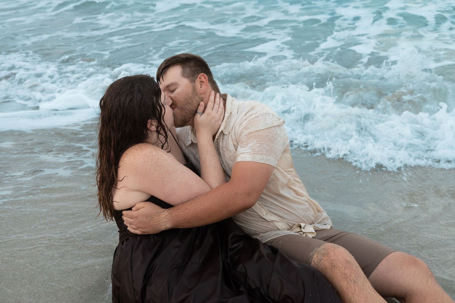 Couple sharing a romantic kiss sitting in ocean waves on the beach.