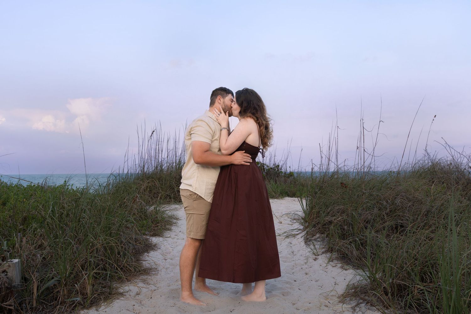 Couple sharing a tender kiss on a sandy beach path surrounded by sea grass.